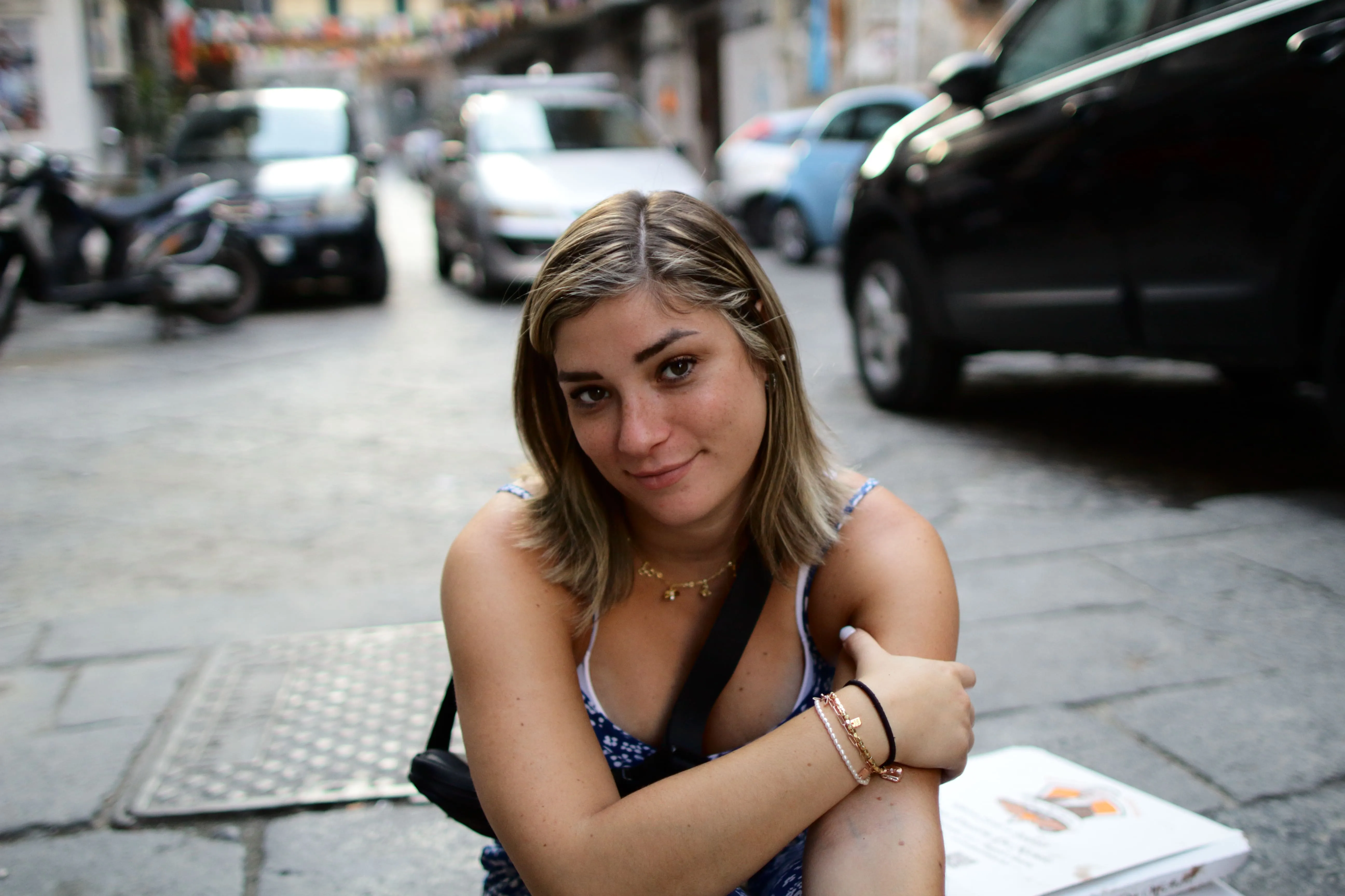 Young woman in blue dress sitting on a street pavement with parked cars in the background.