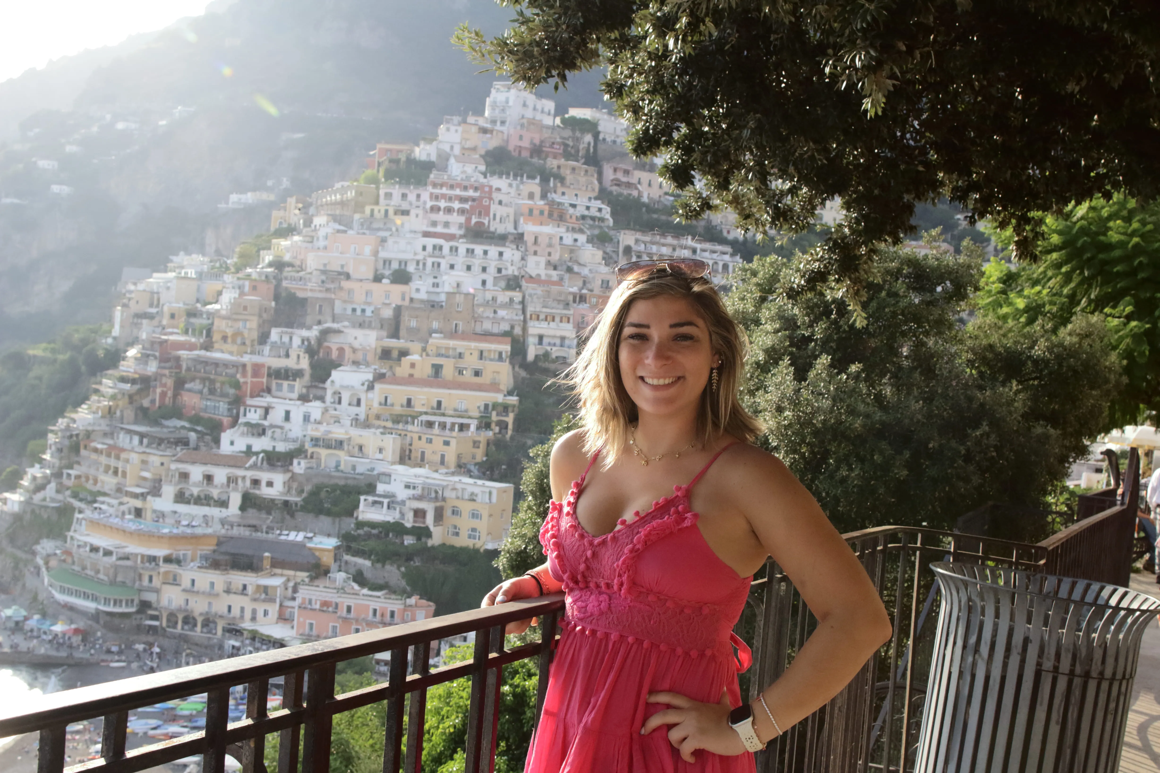 Smiling woman in a pink dress stands by a railing with hillside town buildings in the background.
