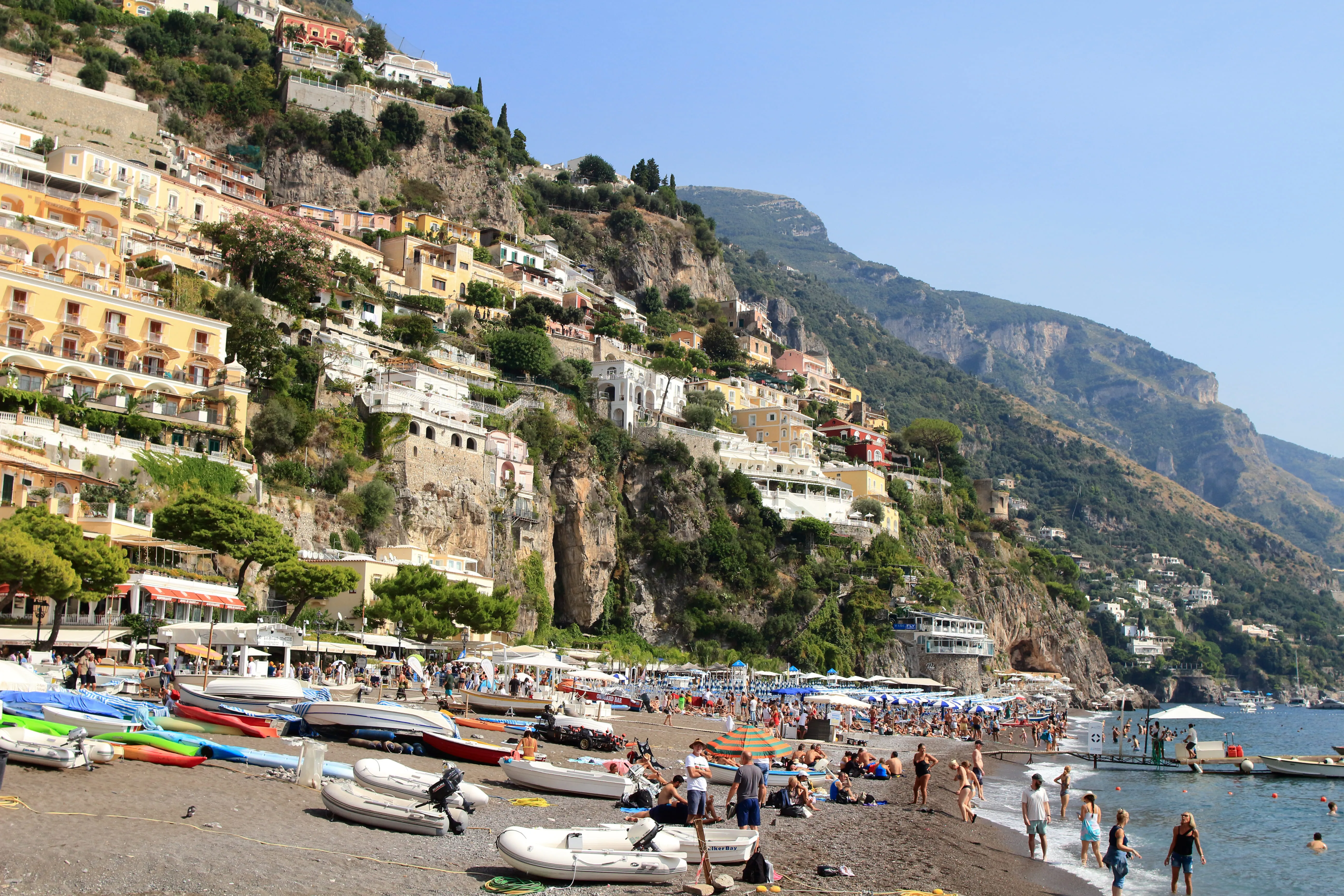 Crowded pebble beach with boats and colorful cliffside houses under clear blue sky.