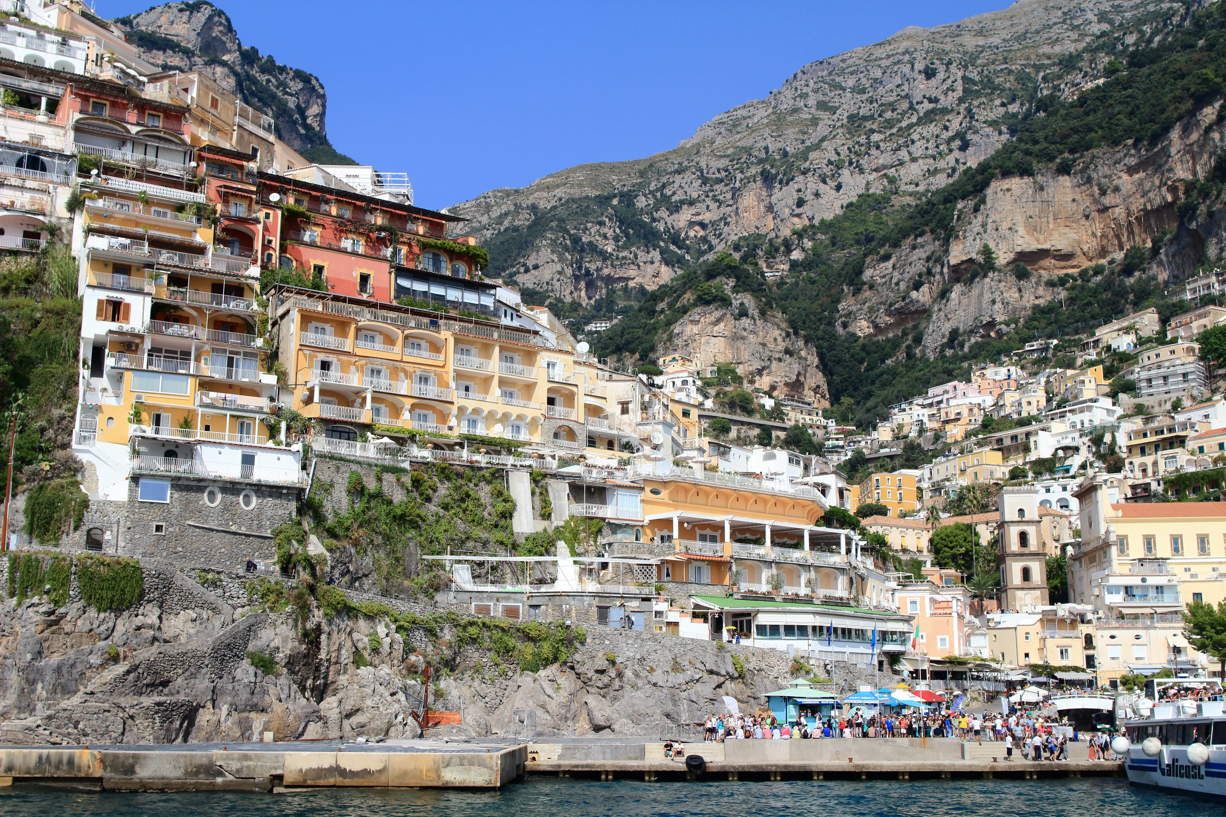 Colorful buildings on rocky cliffs with a crowd and boat near a Mediterranean coastline under clear sky.