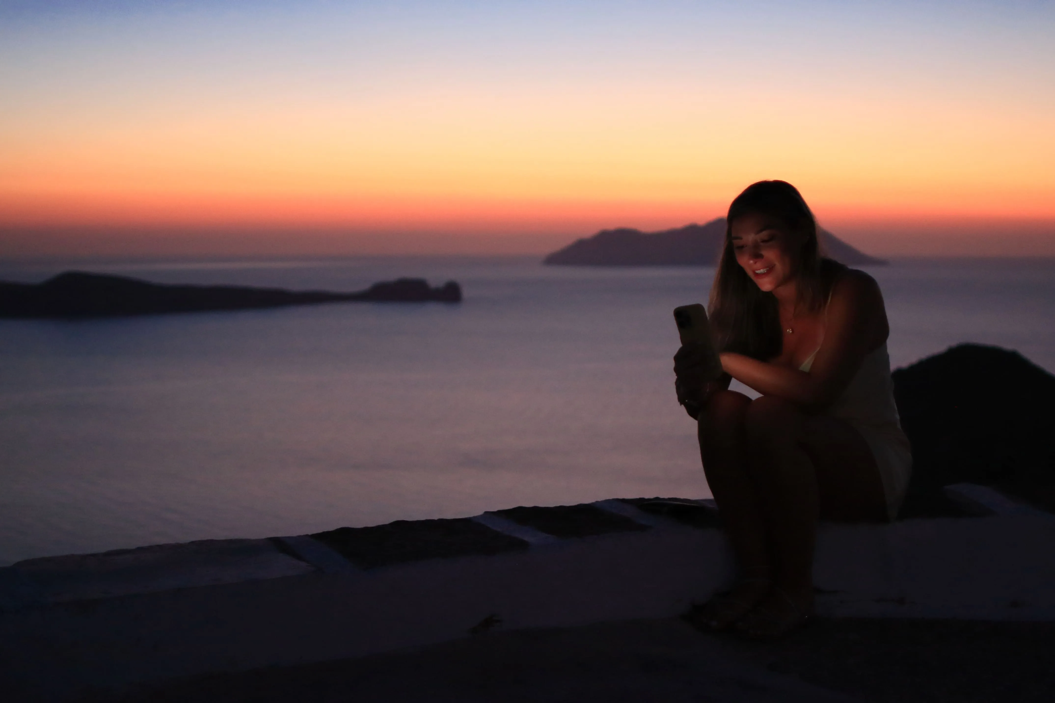 Young woman sitting by the sea at sunset, smiling while looking at her phone.