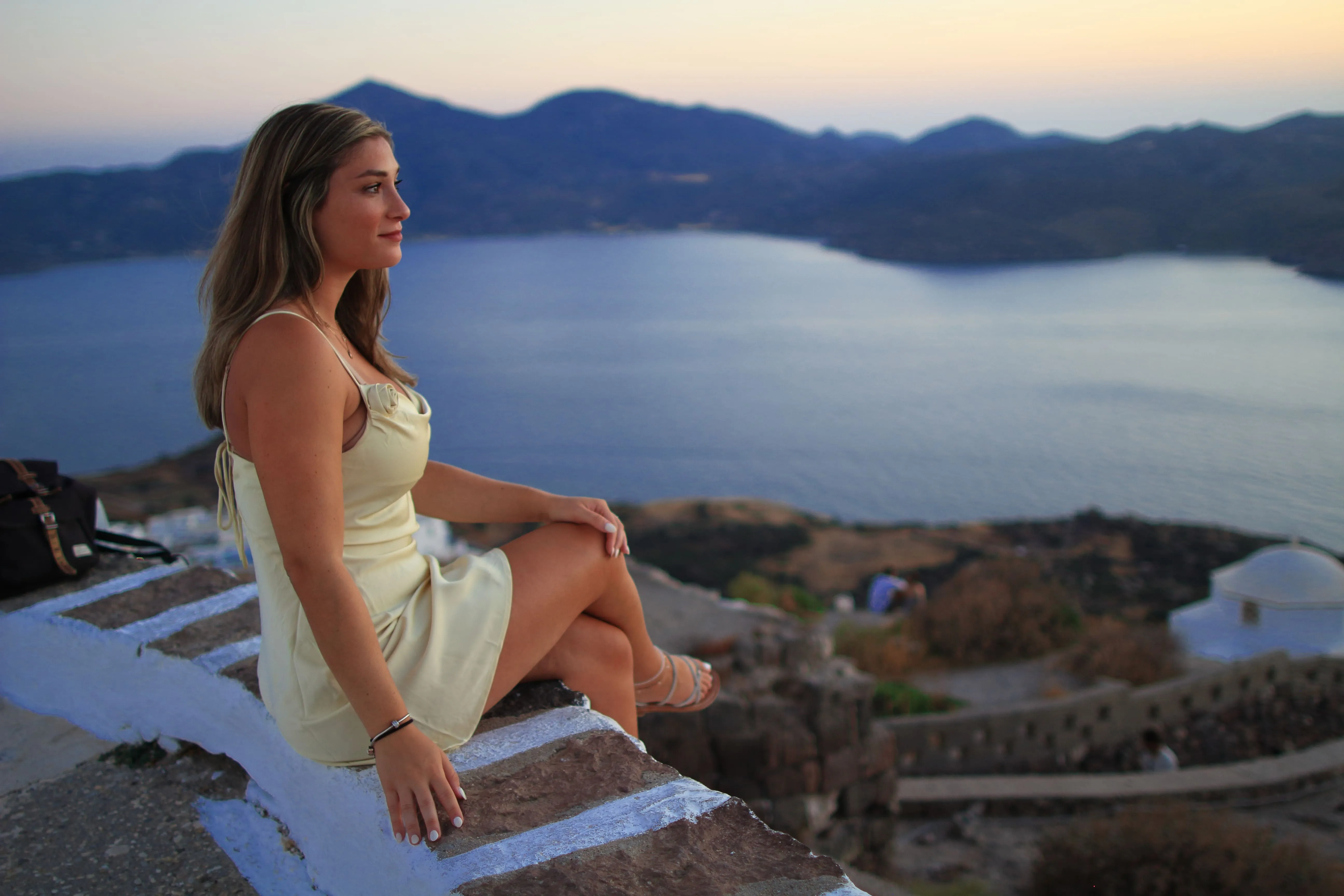 Woman in yellow dress sitting on stone wall overlooking sea and mountains at sunset.