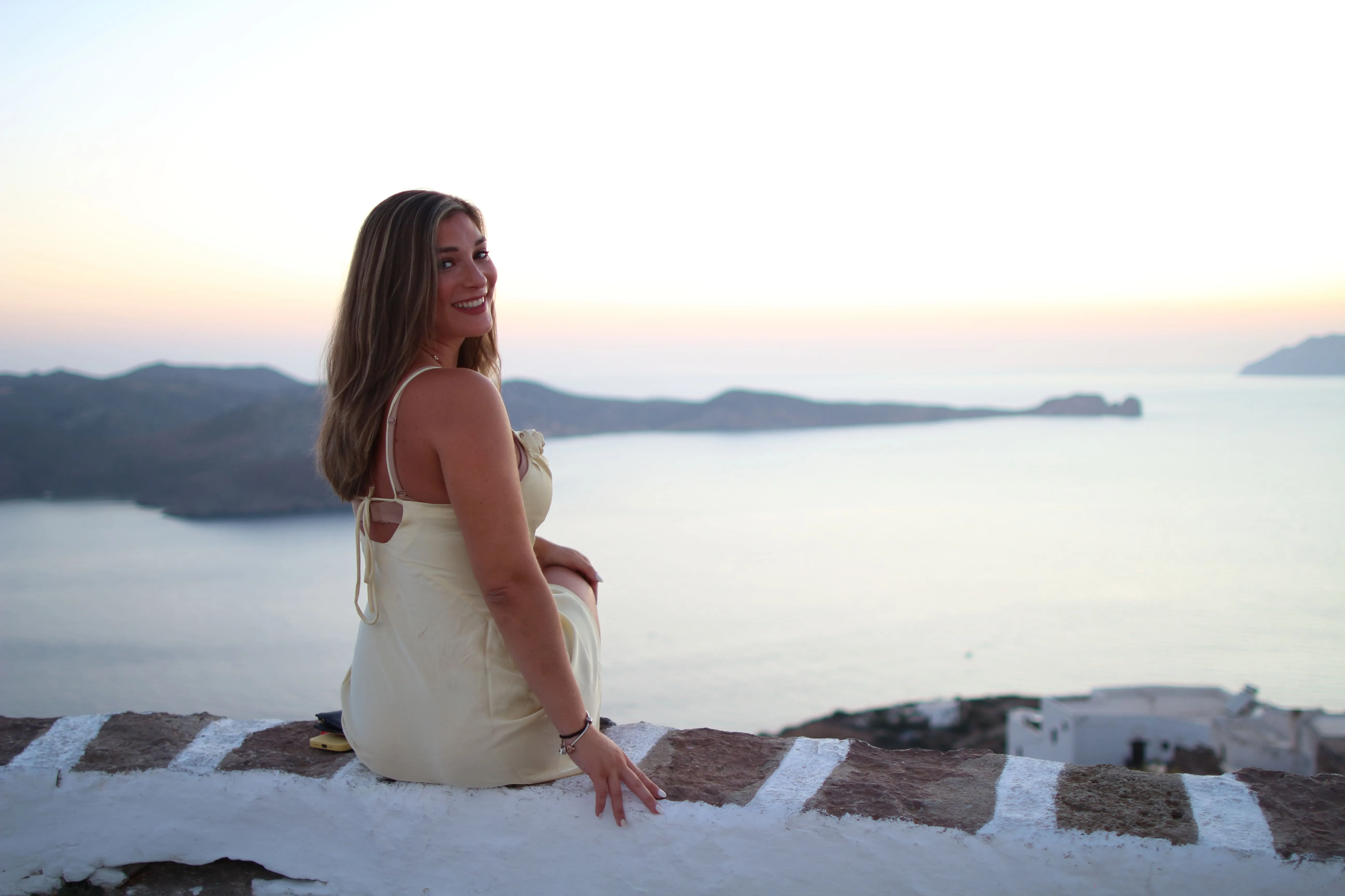 Woman in a light dress sitting on stone wall smiling with sea and islands at sunset in the background.