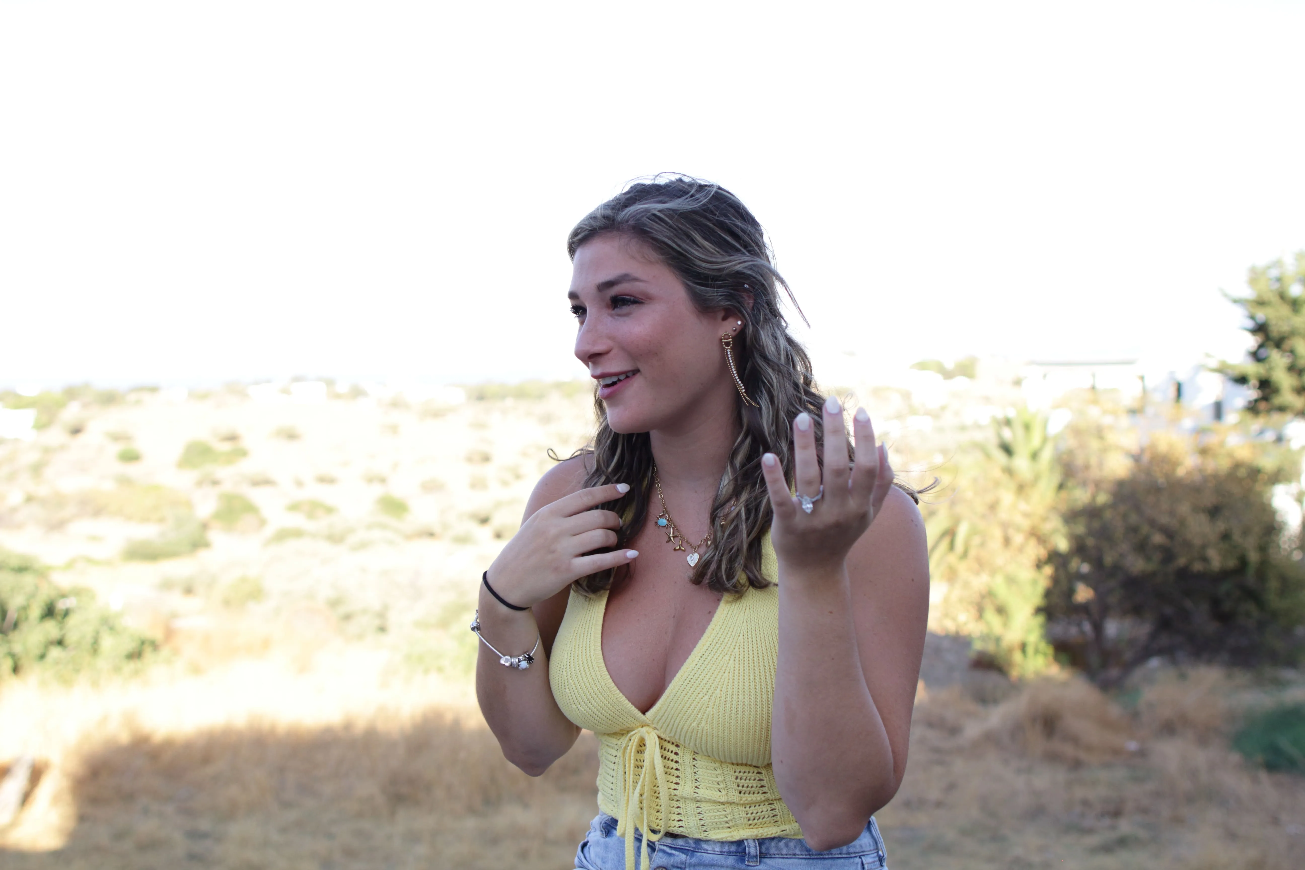 Young woman smiling outdoors wearing a yellow sleeveless top and denim shorts with jewelry.