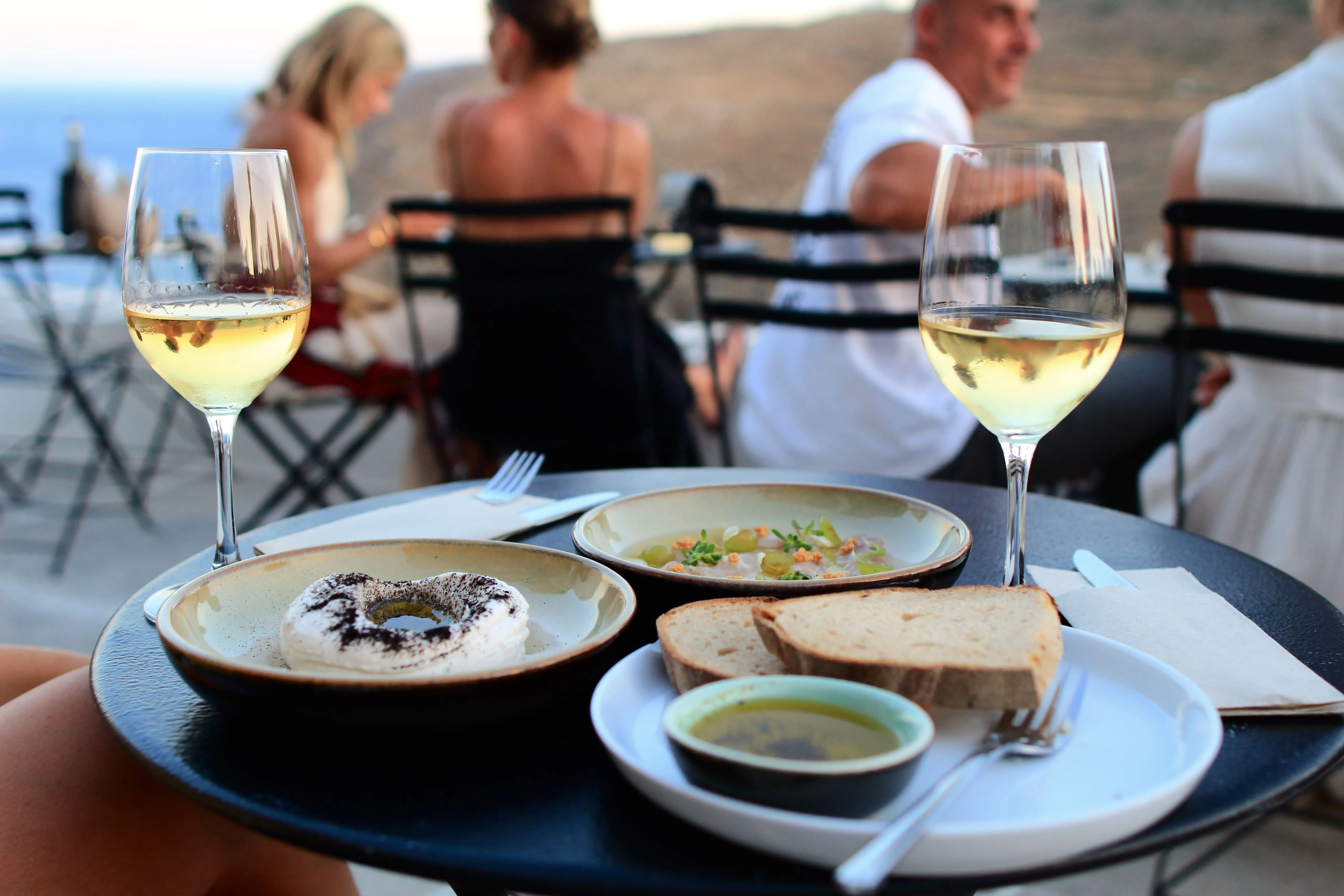 Two glasses of white wine with plates of bread, dip, and dessert on a black table outdoors.