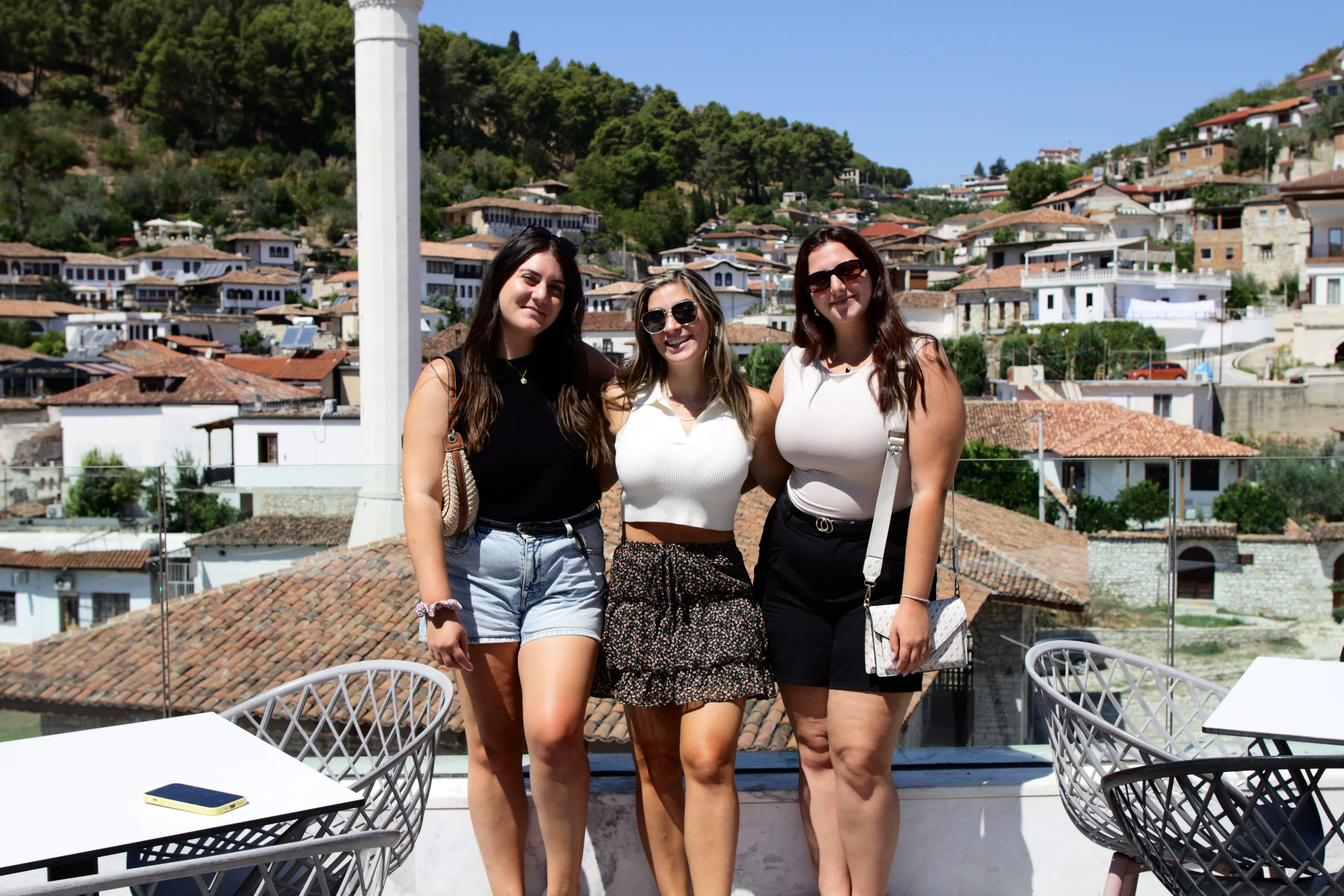 Three women in summer clothes standing arm in arm on a terrace with village rooftops and a minaret behind.