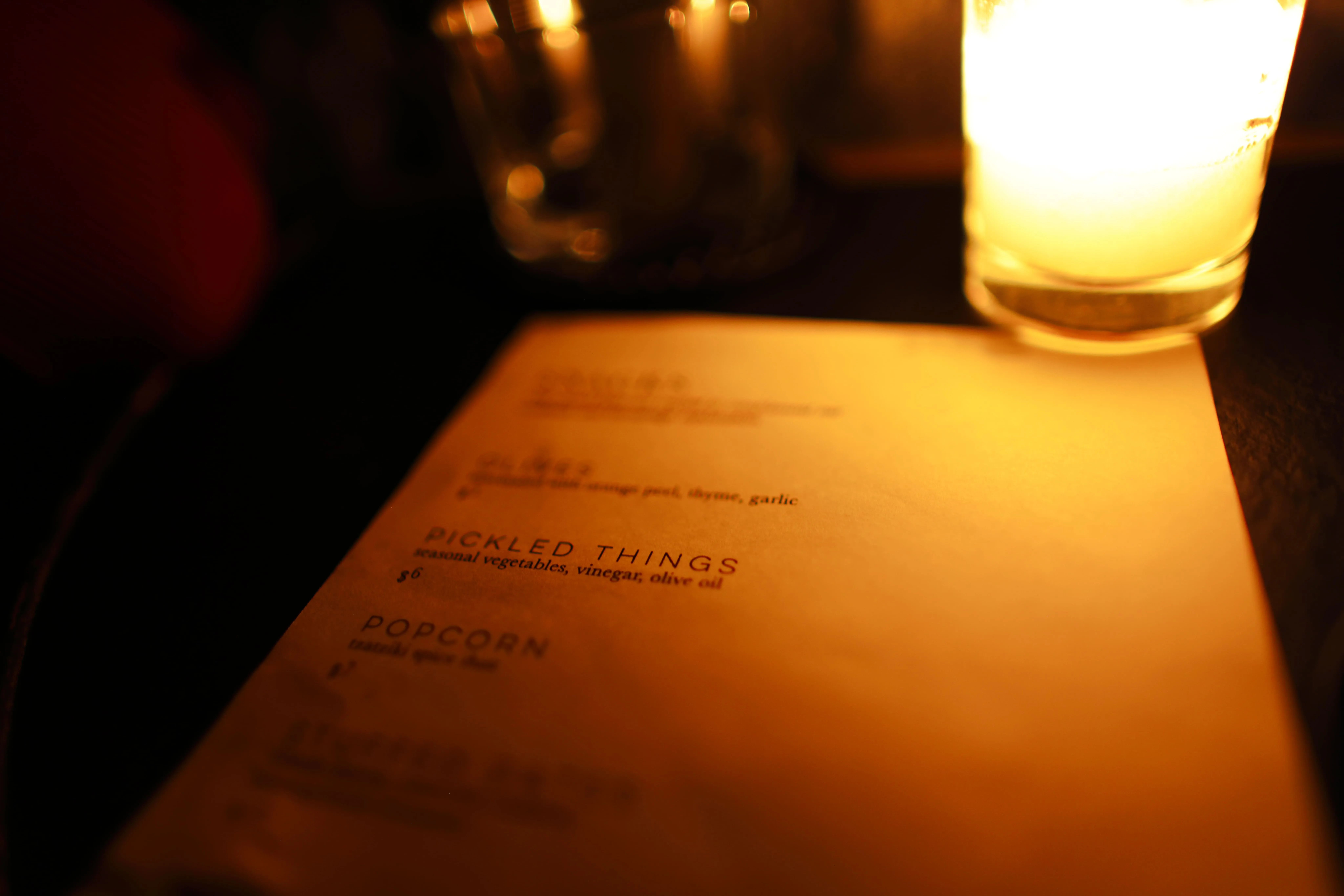 Dimly lit menu on a table showing items like pickled things, olives, and popcorn with a glowing candle nearby.