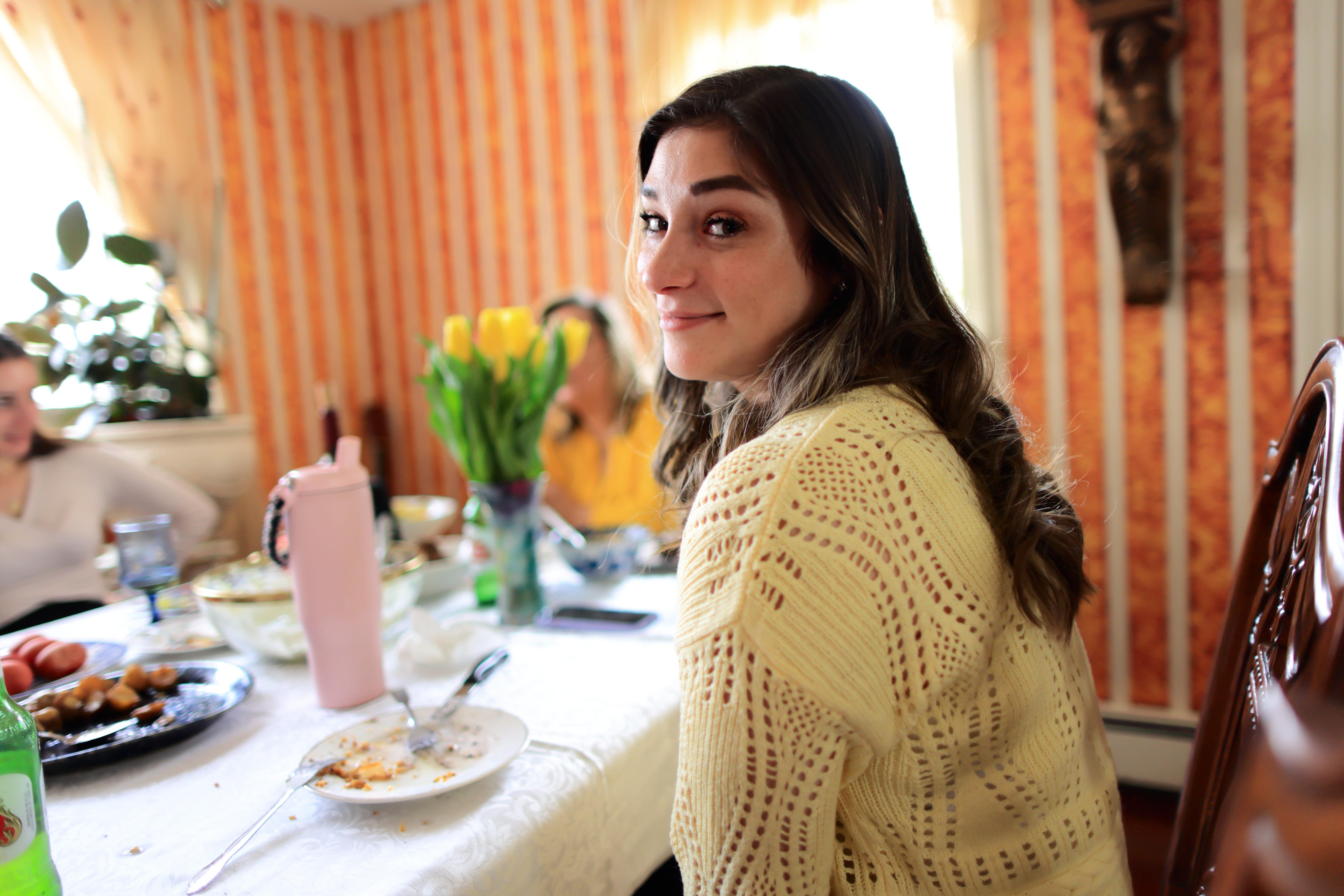Woman in a yellow sweater smiles at camera at a dining table with food and yellow tulips.