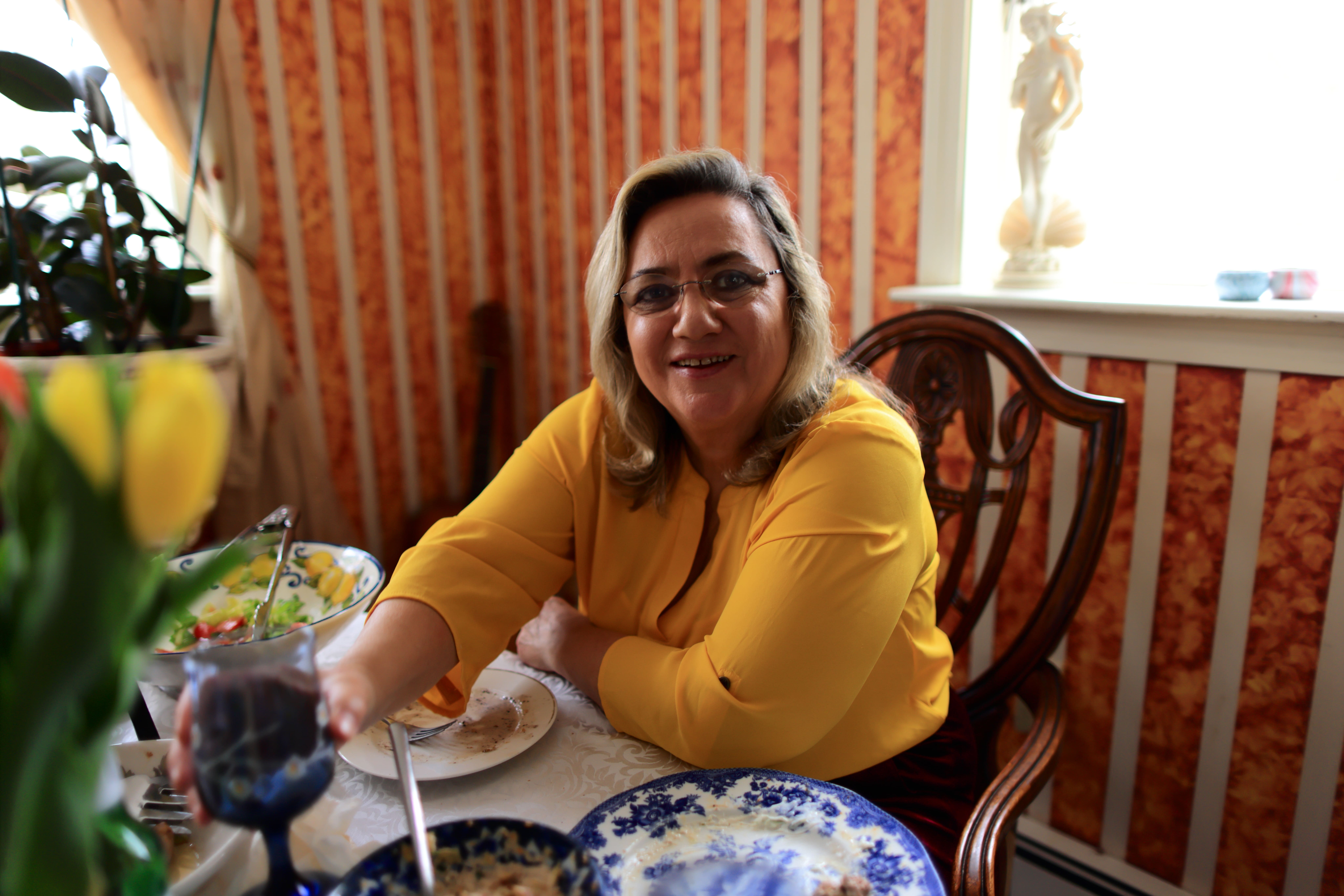 Smiling woman in yellow blouse sitting at a table with plates and reaching for a blue glass.
