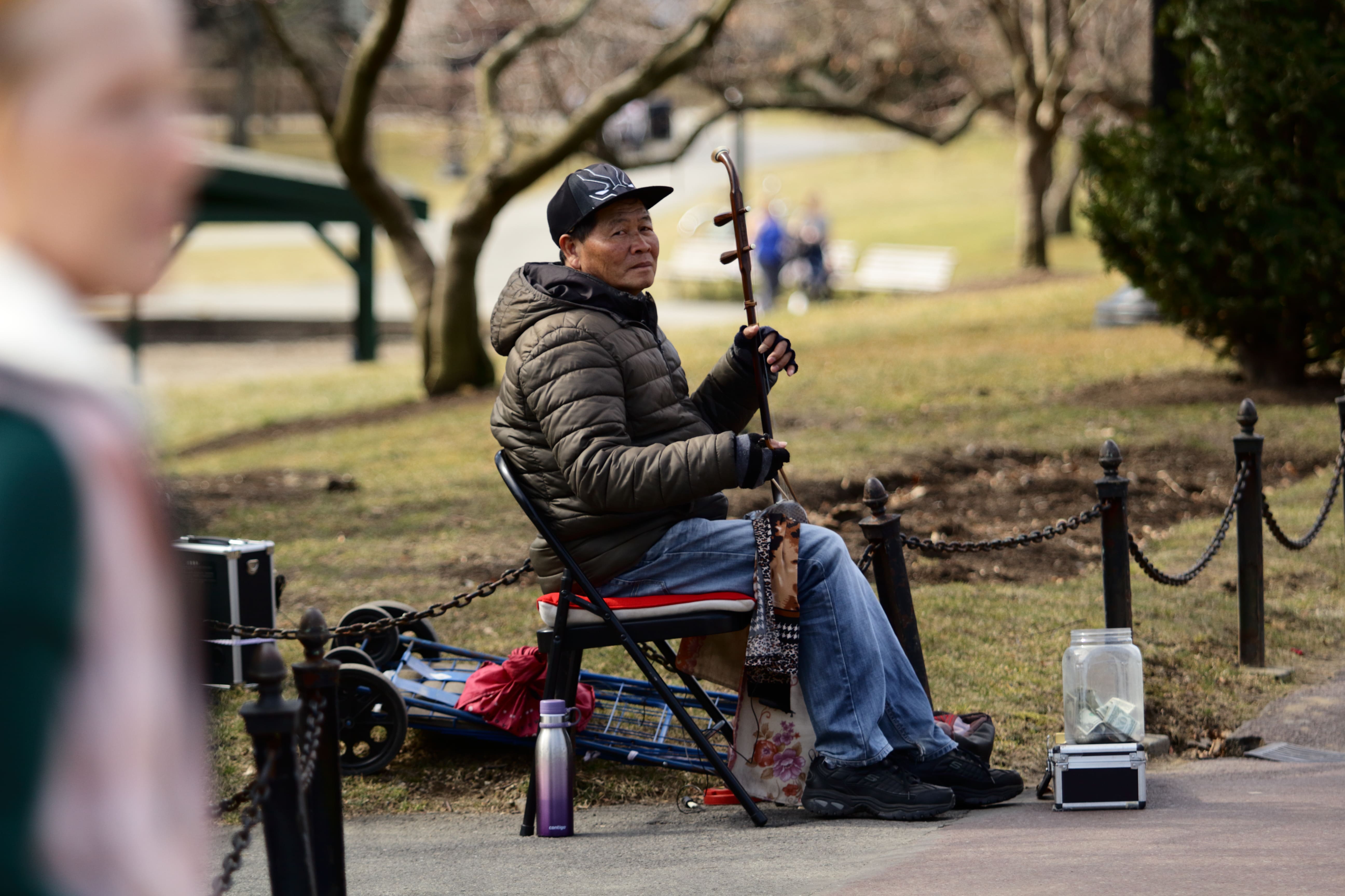 Man in a jacket playing a string instrument while seated on a folding chair in a park.