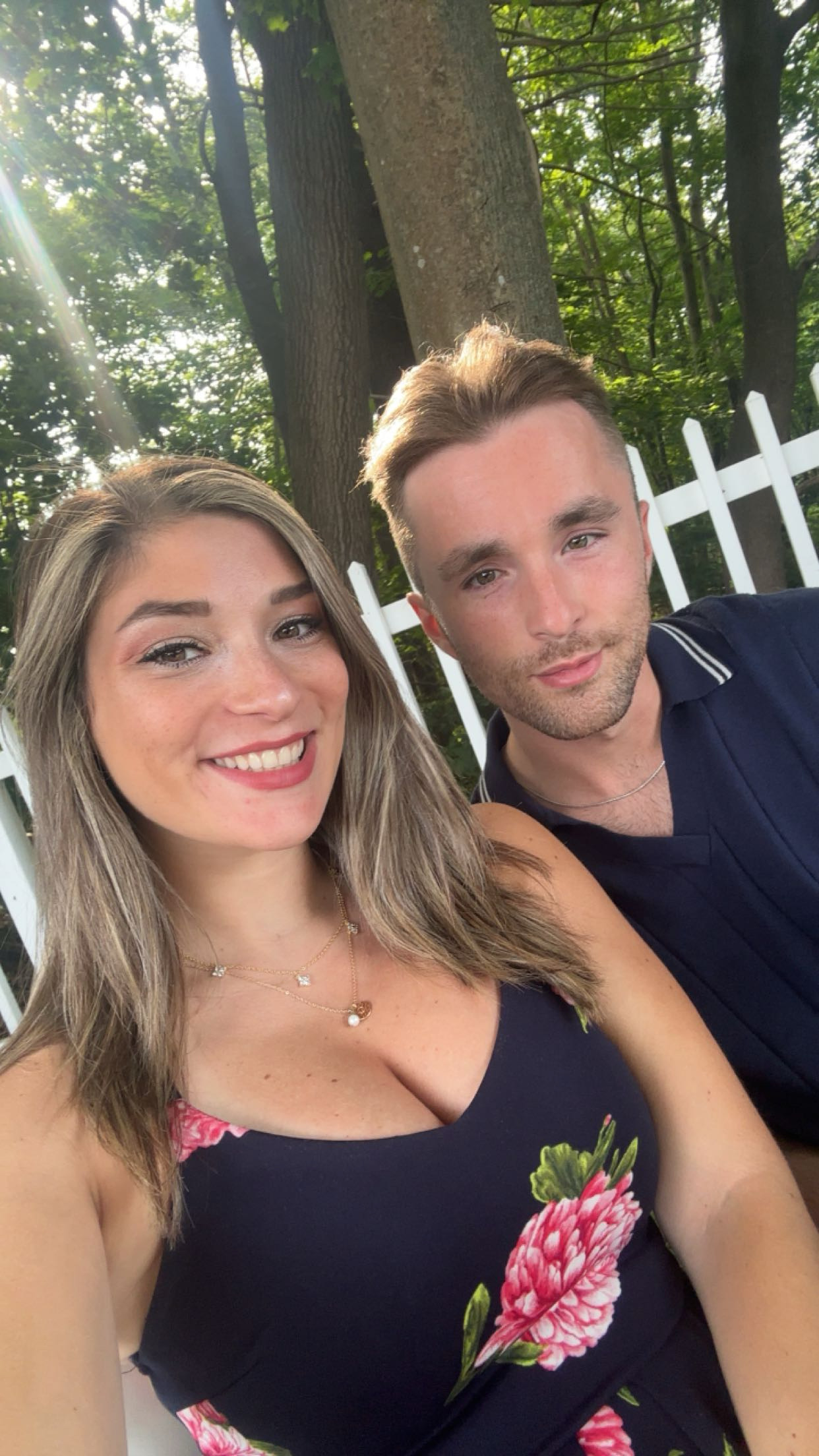 Smiling woman in floral dress and man in navy shirt sitting outdoors by white fence and trees.