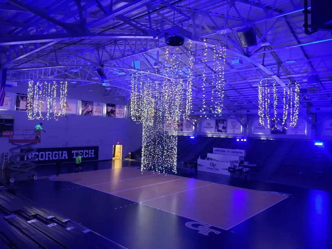 Indoor gymnasium with hanging decorative string lights illuminated in blue and white above a sports court with Georgia Tech logos.