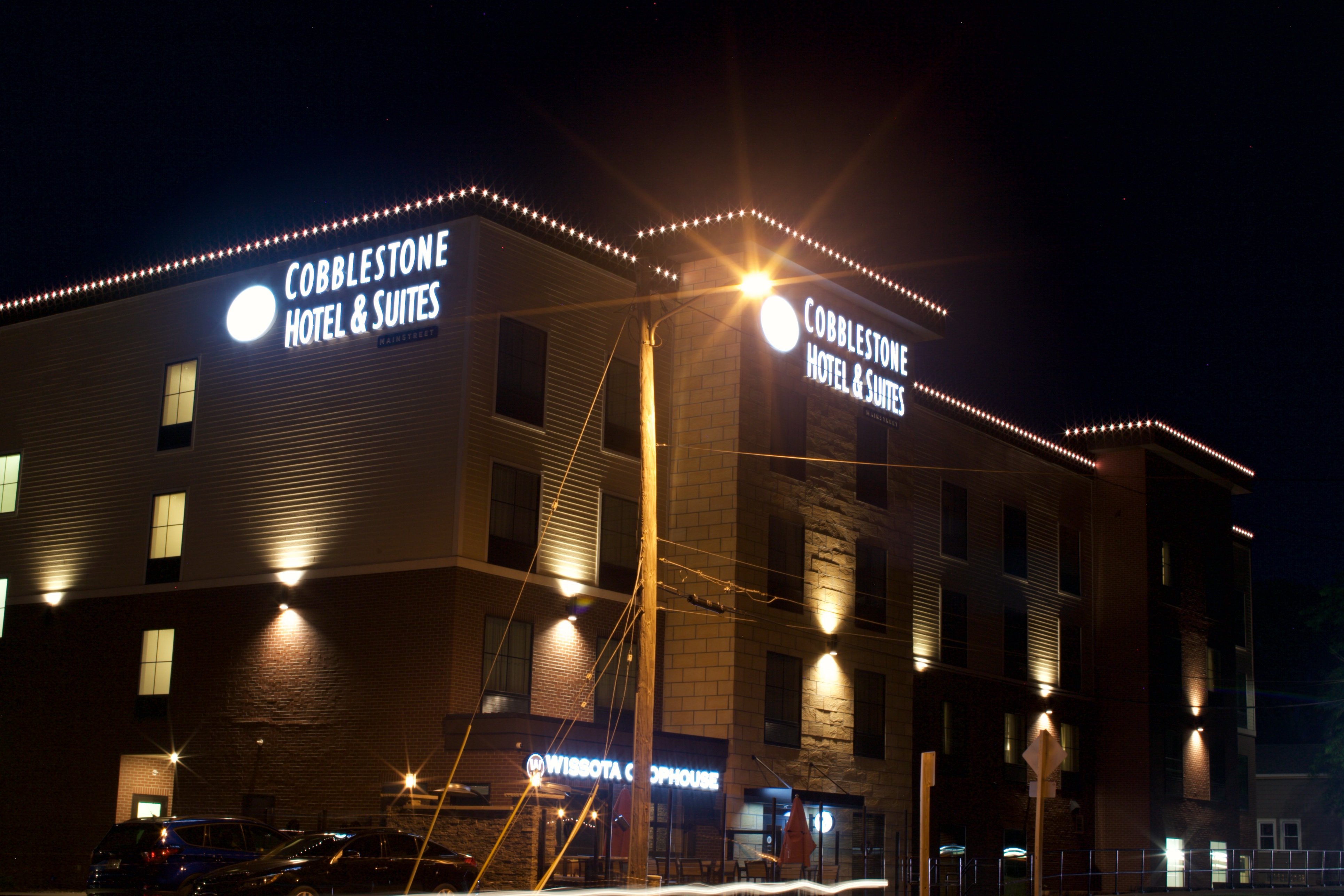 Exterior of Cobblestone Hotel & Suites at night with illuminated signage on the building corners, outdoor lighting, and parked cars in front.