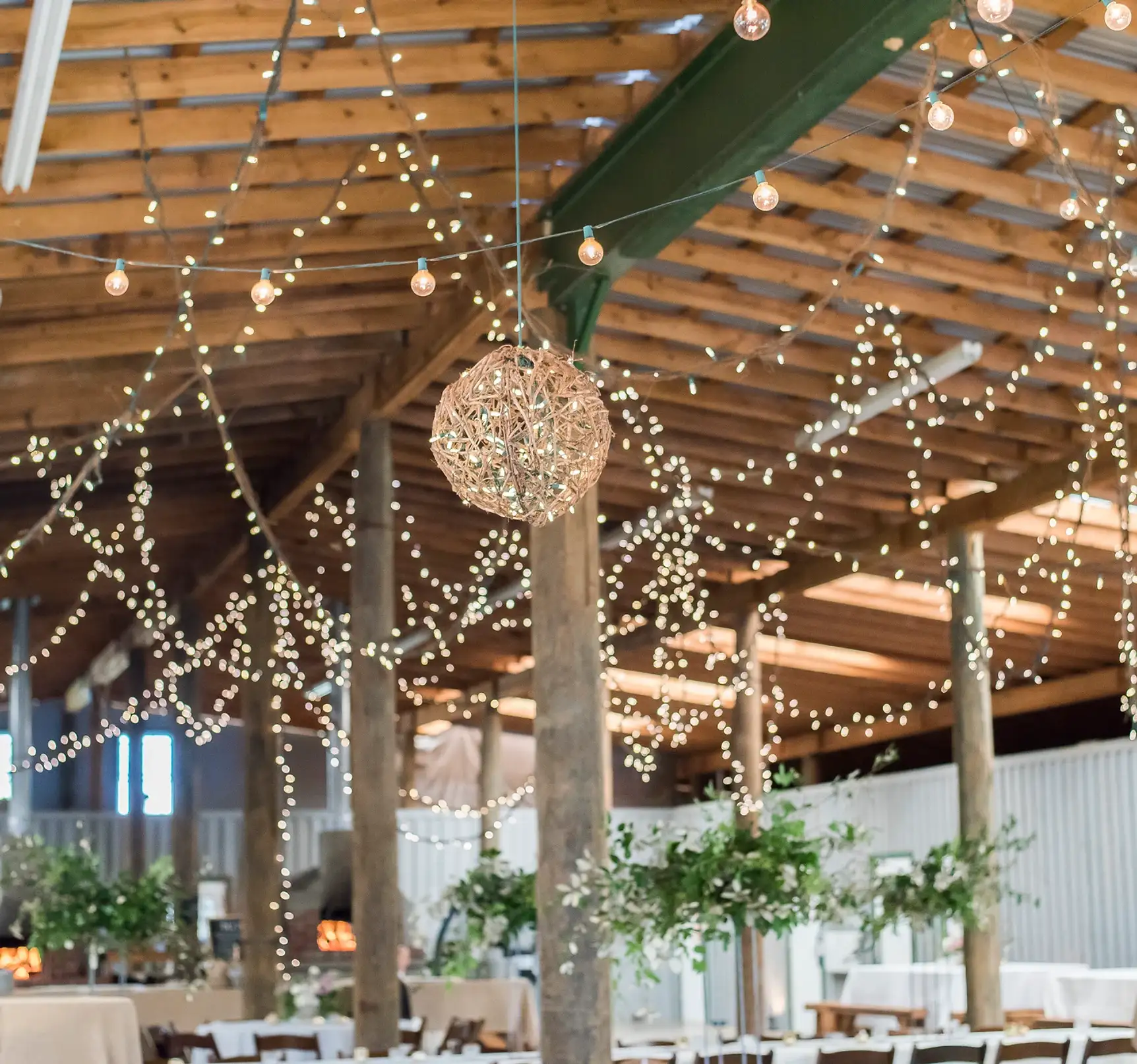 Rustic barn interior with wooden beams and string lights hanging above tables with floral centerpieces.