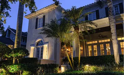 Well-lit two-story house with palm trees and bushes in the front yard at dusk.