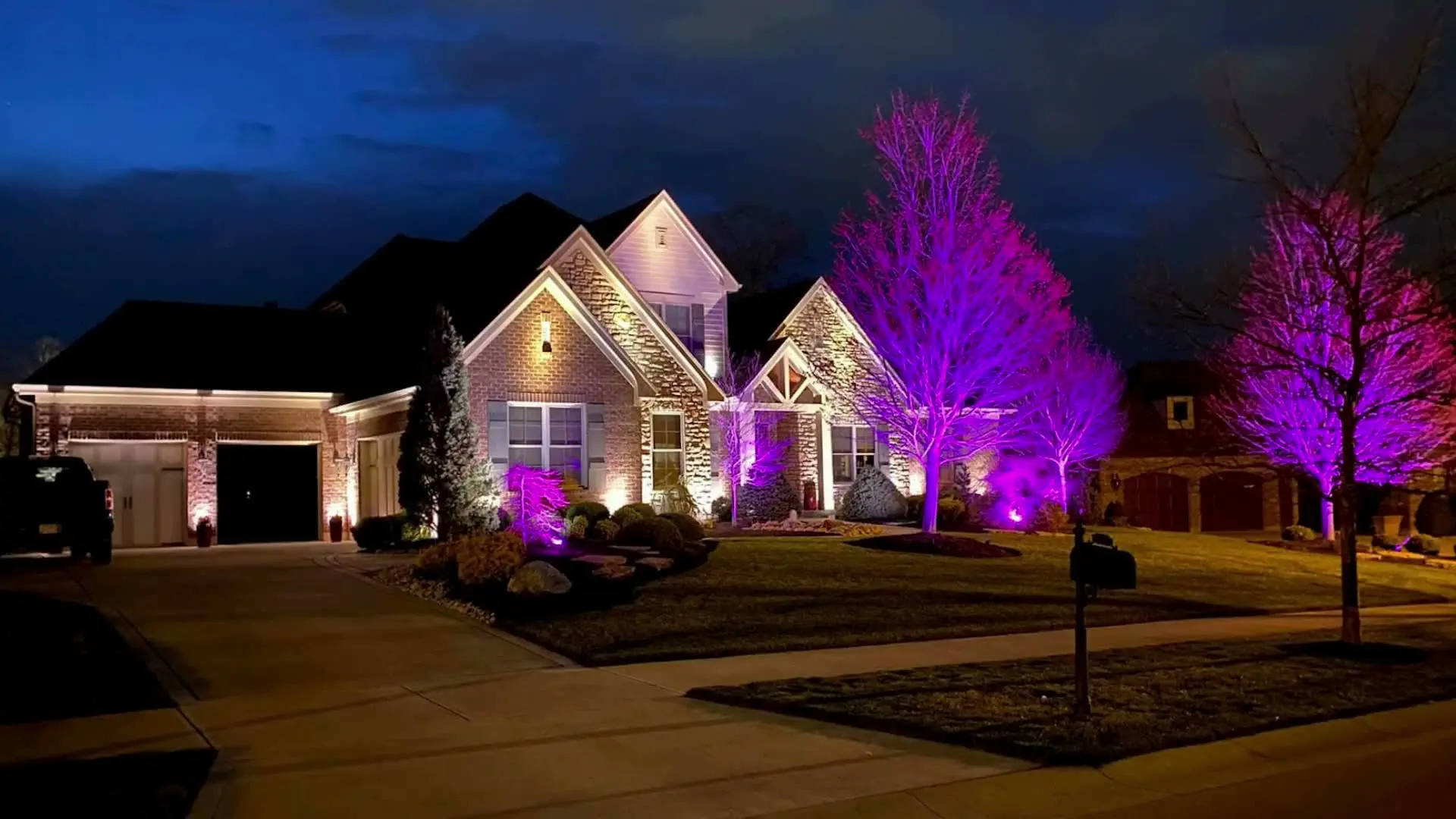Night view of a brick house with purple-lit trees and warm exterior wall lighting.