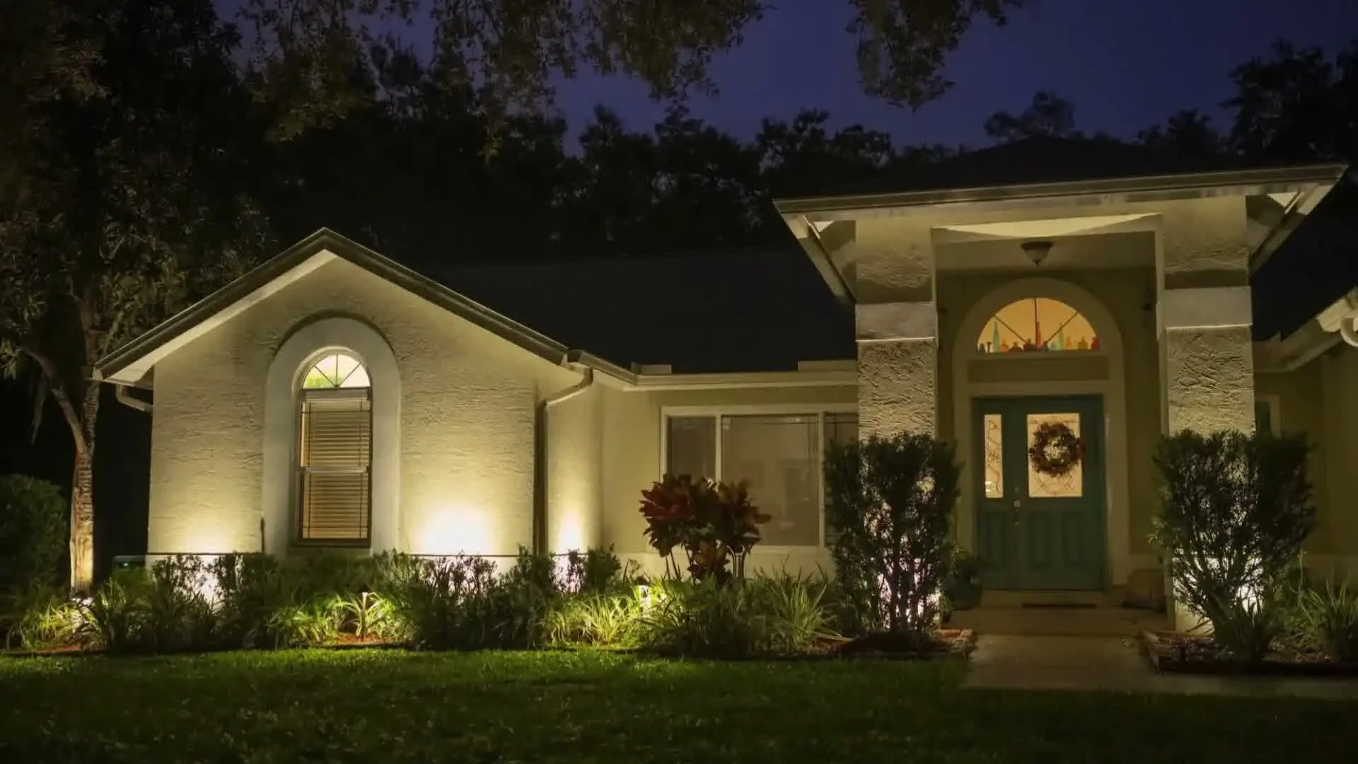 A lit house entrance at night with a wreath on the door and garden plants along the path.