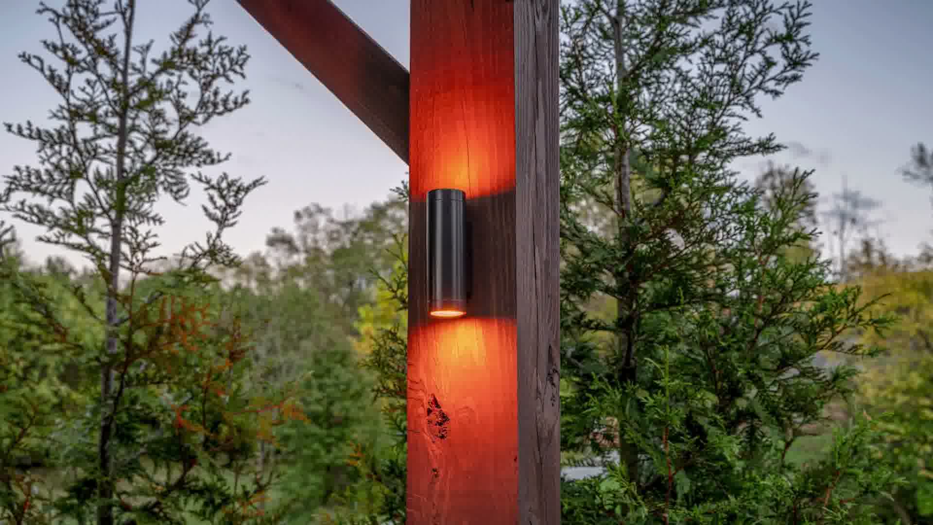 Outdoor wall light fixture glowing orange on a wooden post surrounded by green foliage.