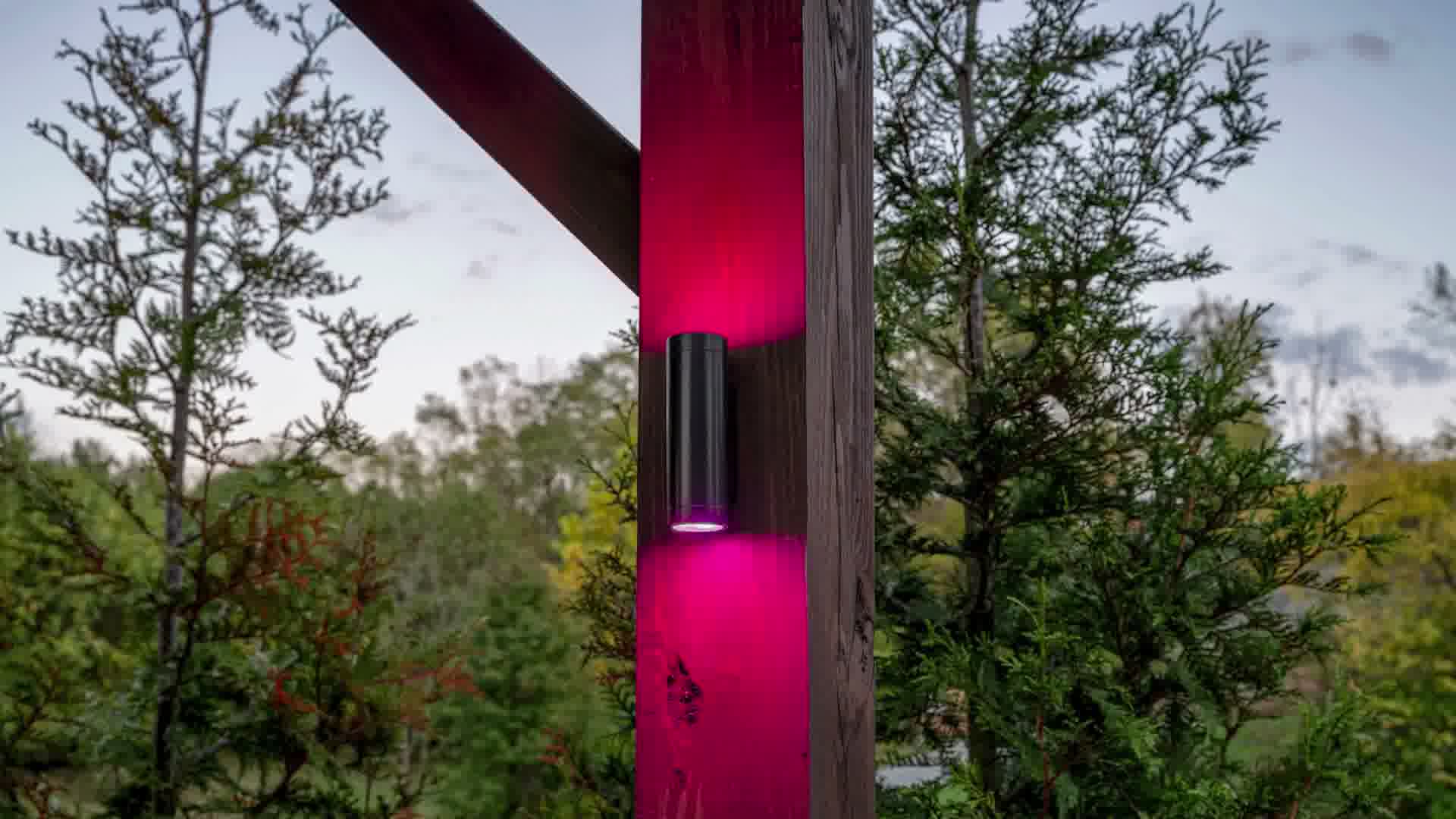 Black outdoor light fixture on wooden post casting pink light at dusk with green trees in background.