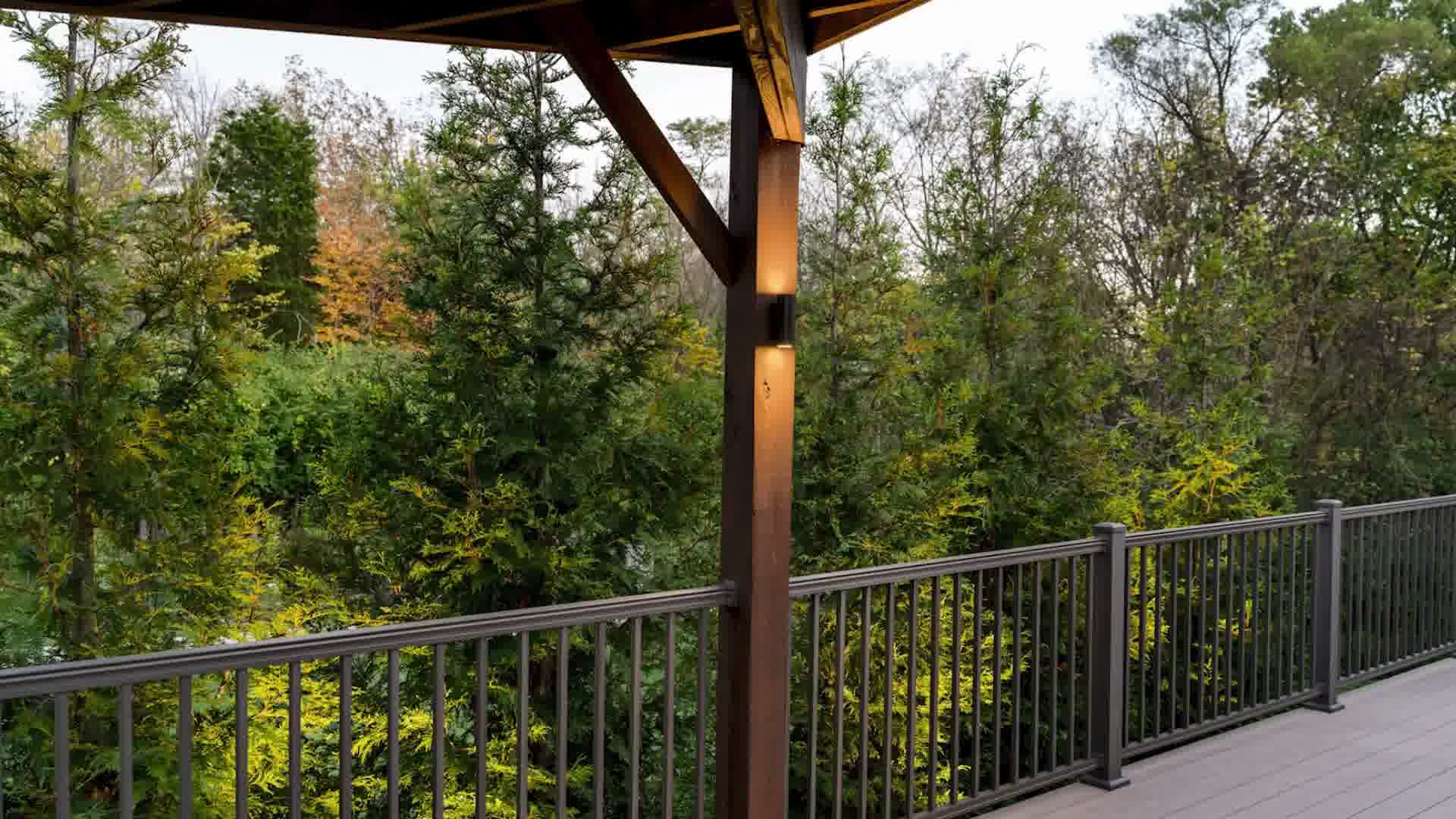 Wooden deck railing with a lit post light and dense green trees in the background.
