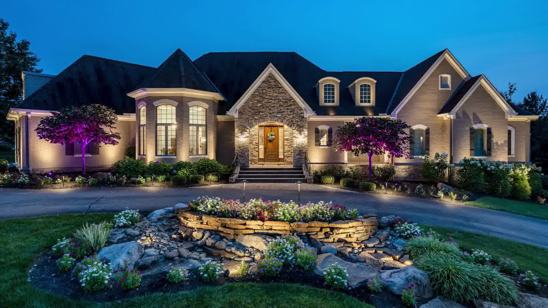 Large house lit with warm lights, purple-lit trees, stone entrance, and flower garden with rocks in front.