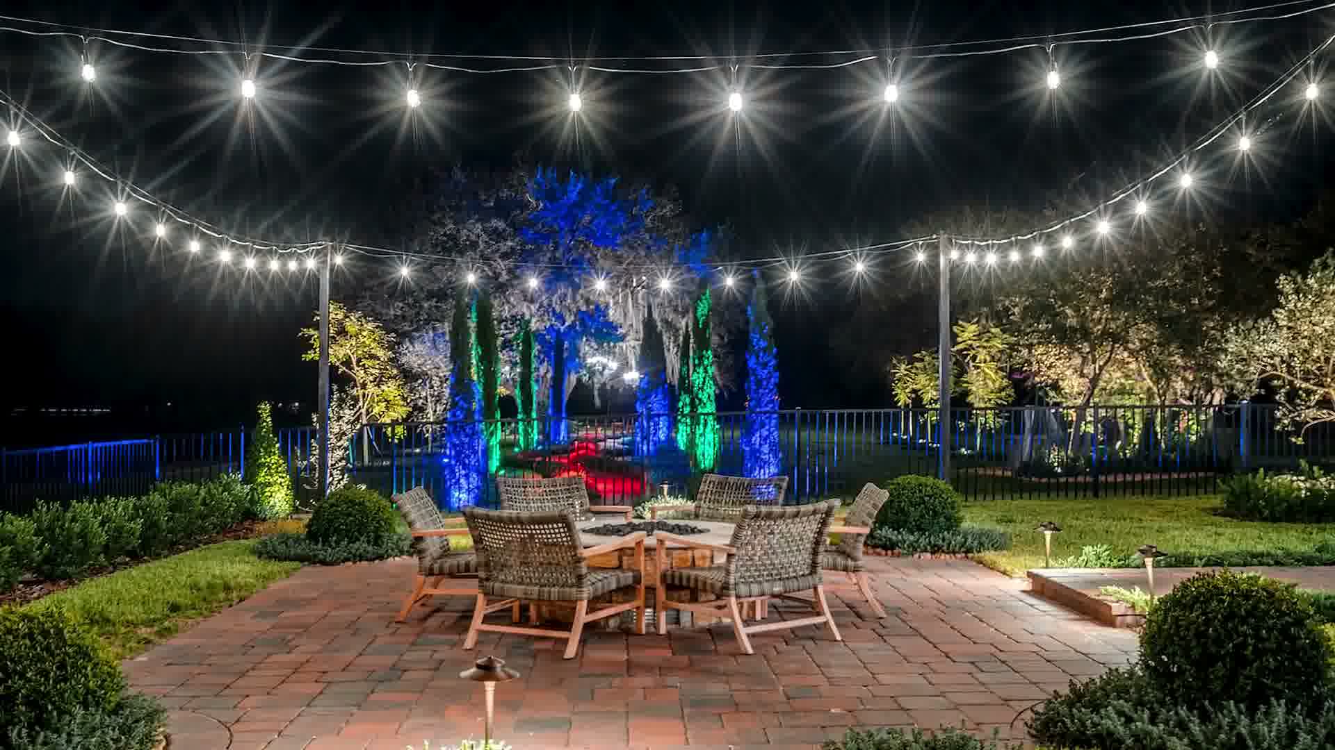 Outdoor patio at night with string lights, six woven chairs around a fire pit, and colorful lit trees.