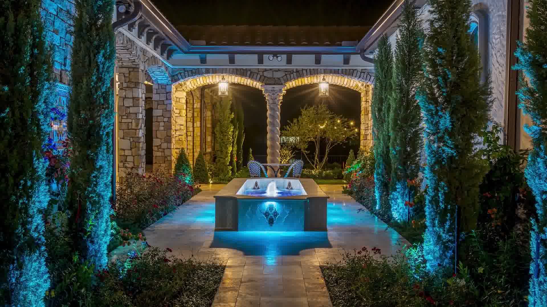 Outdoor patio at night with fountain, blue lighting, stone arches, greenery, and two chairs with a small table.