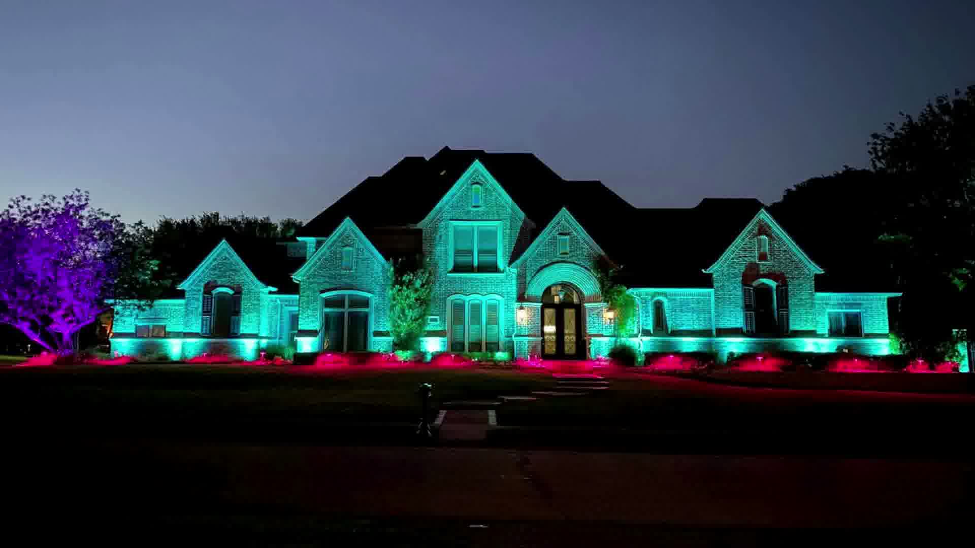 Large house at night lit with turquoise and pink exterior lighting with purple-lit tree on left.