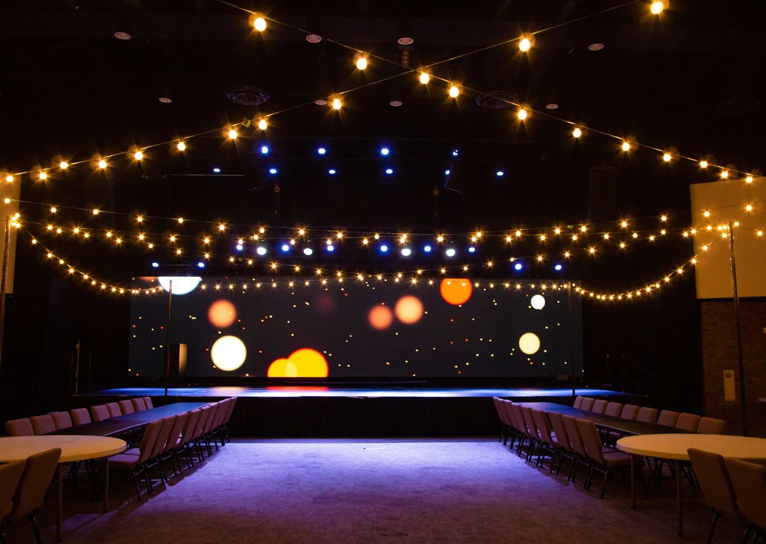 Indoor auditorium with string lights overhead, long tables with chairs, and a stage with colorful lights.
