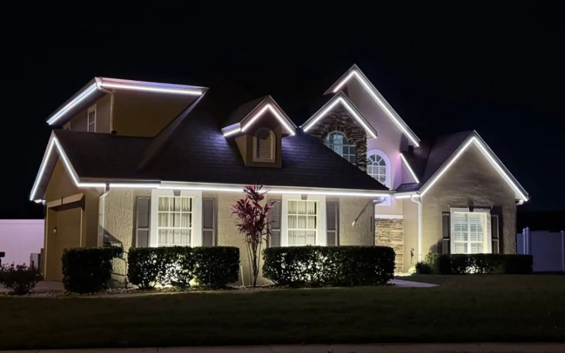 Two-story house at night with white LED trim lights outlining the roof and illuminated bushes.