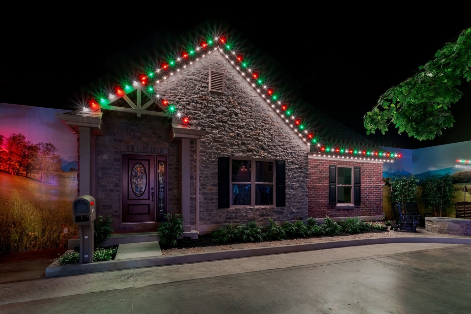 Stone and brick house at night with red, green, and white Christmas lights along the roofline, a wooden front door with decorative glass panels, a mailbox near the walkway, shrubbery along the foundation, and a mural of a landscape with trees and mountains in the background.