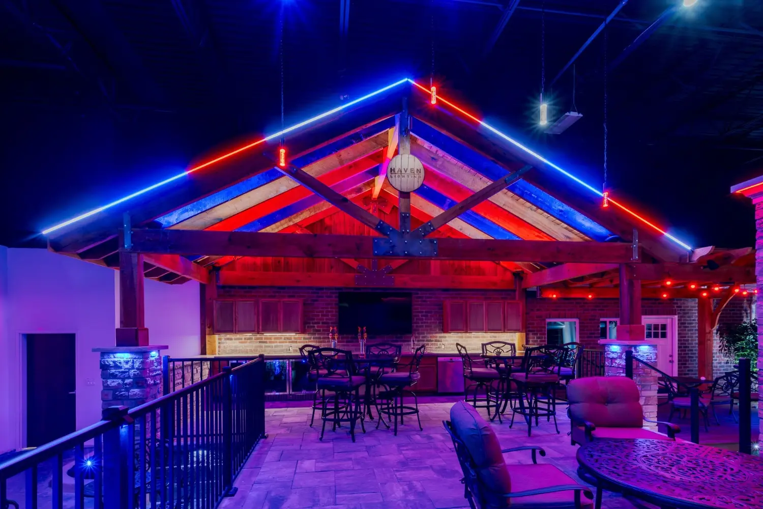 Outdoor patio area at night featuring a wooden pergola with pitched roof highlighted by red and blue LED strip lights, multiple high tables with chairs, cushioned seats, and railings, creating a vibrant and inviting atmosphere.