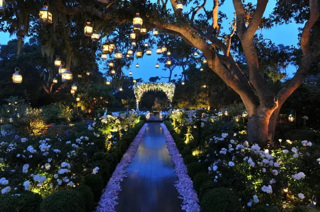 Evening garden pathway lined with white flowers and hanging lanterns leading to a floral archway.