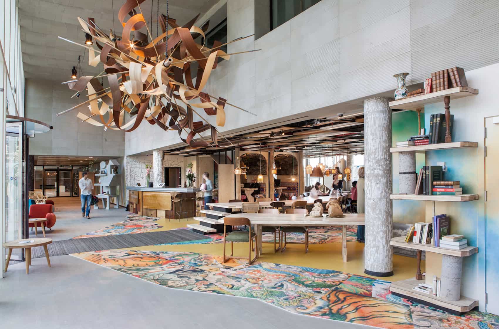 Modern office lobby with colorful patterned carpet, large wooden chandelier, bookshelves, and people working in the background.