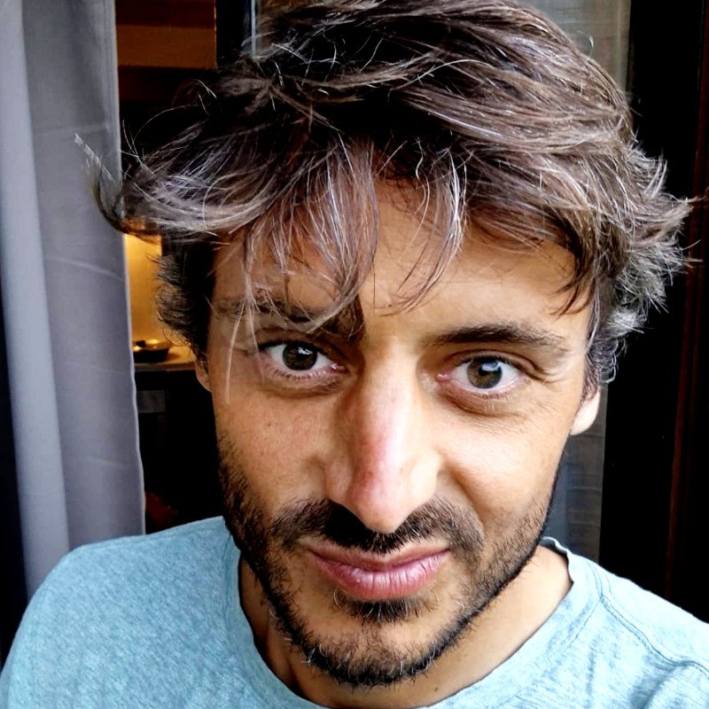 Close-up of a man with brown hair and a beard, wearing a light blue shirt, looking directly at the camera.