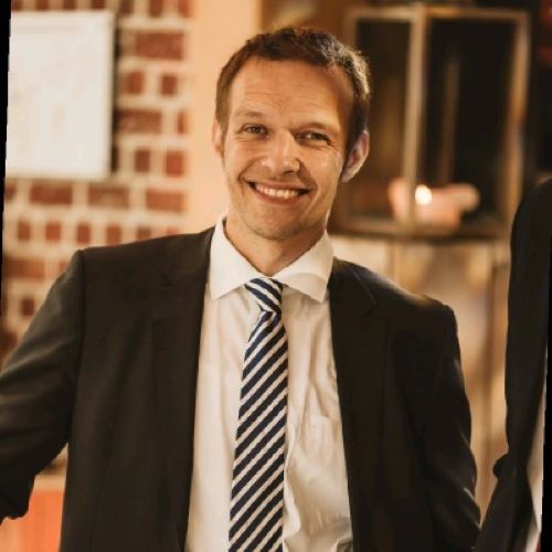 Smiling man wearing a dark suit, white shirt, and striped tie standing indoors.