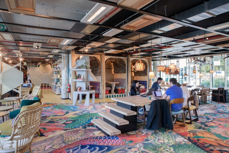 Modern coworking space with colorful patterned carpet, wooden tables, booth seating with pendant lights, and people working on laptops.