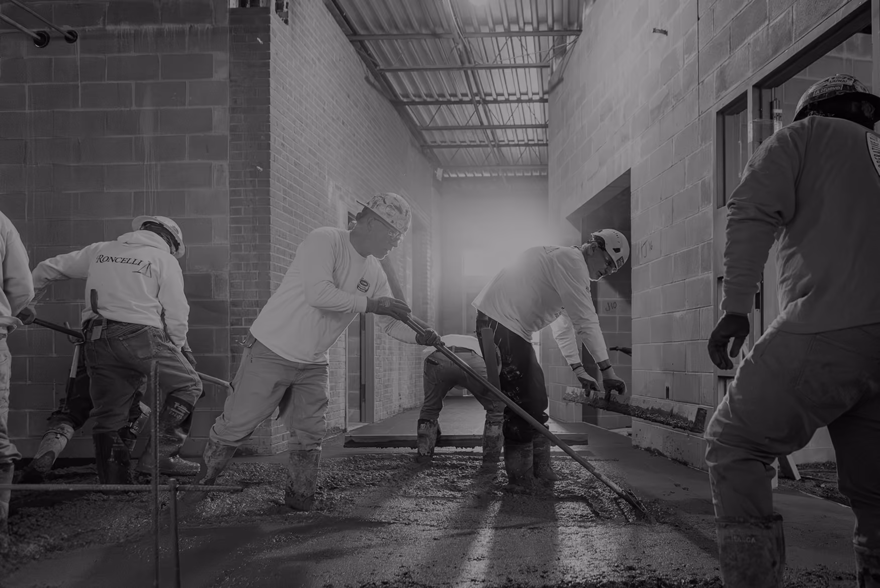 Construction workers wearing hard hats and boots spreading wet concrete on a floor inside a building under construction.