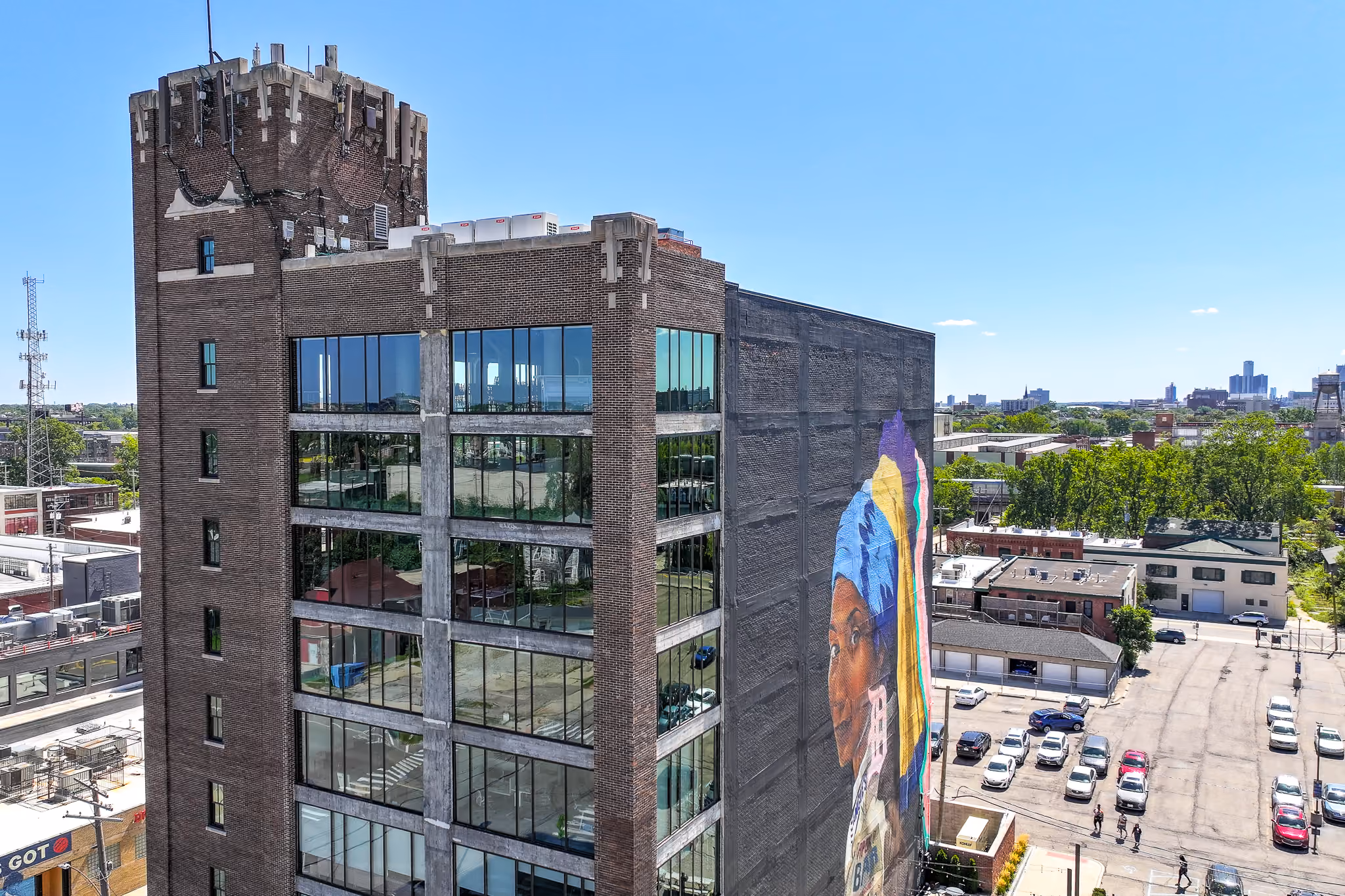 A tall brick building with large glass windows and a mural of a woman wearing a blue headscarf painted on its side, surrounded by a parking lot and other buildings under a clear blue sky.