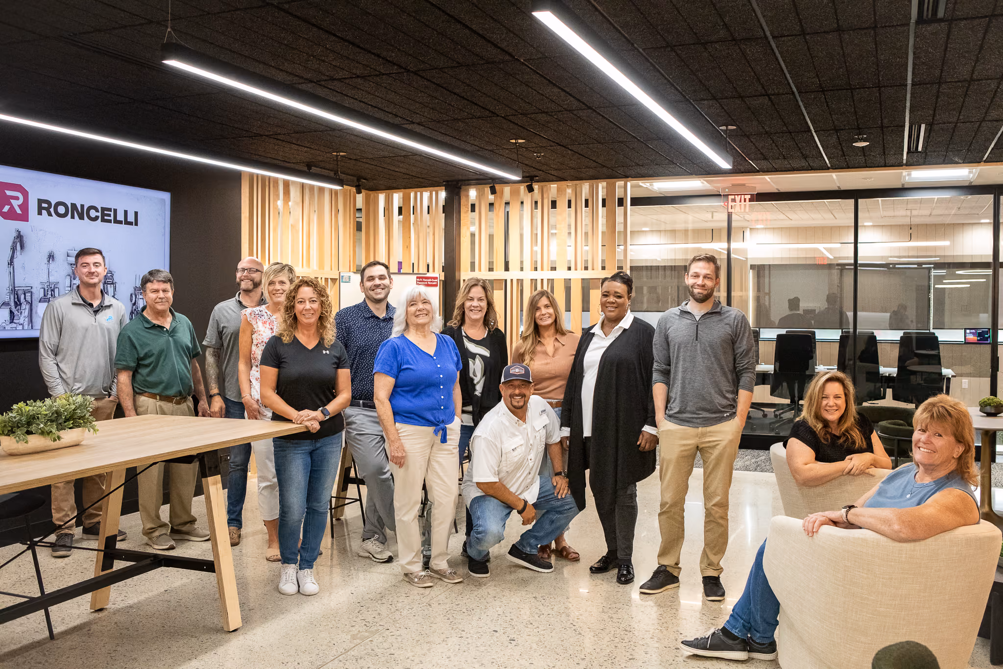 A diverse group of thirteen office team members posing together in a modern workspace with wooden accents and glass walls, some standing, one kneeling, and two seated on white chairs, with a screen displaying the logo 'Roncellow' on the left.