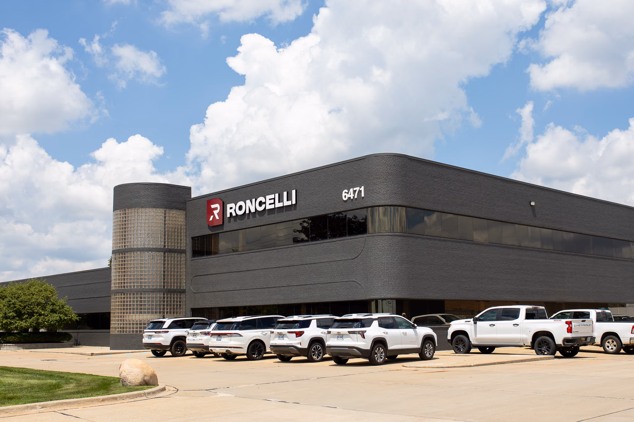 Modern two-story office building with the name Roncelli and number 6471 on its facade under a partly cloudy sky, with several white vehicles parked in front.