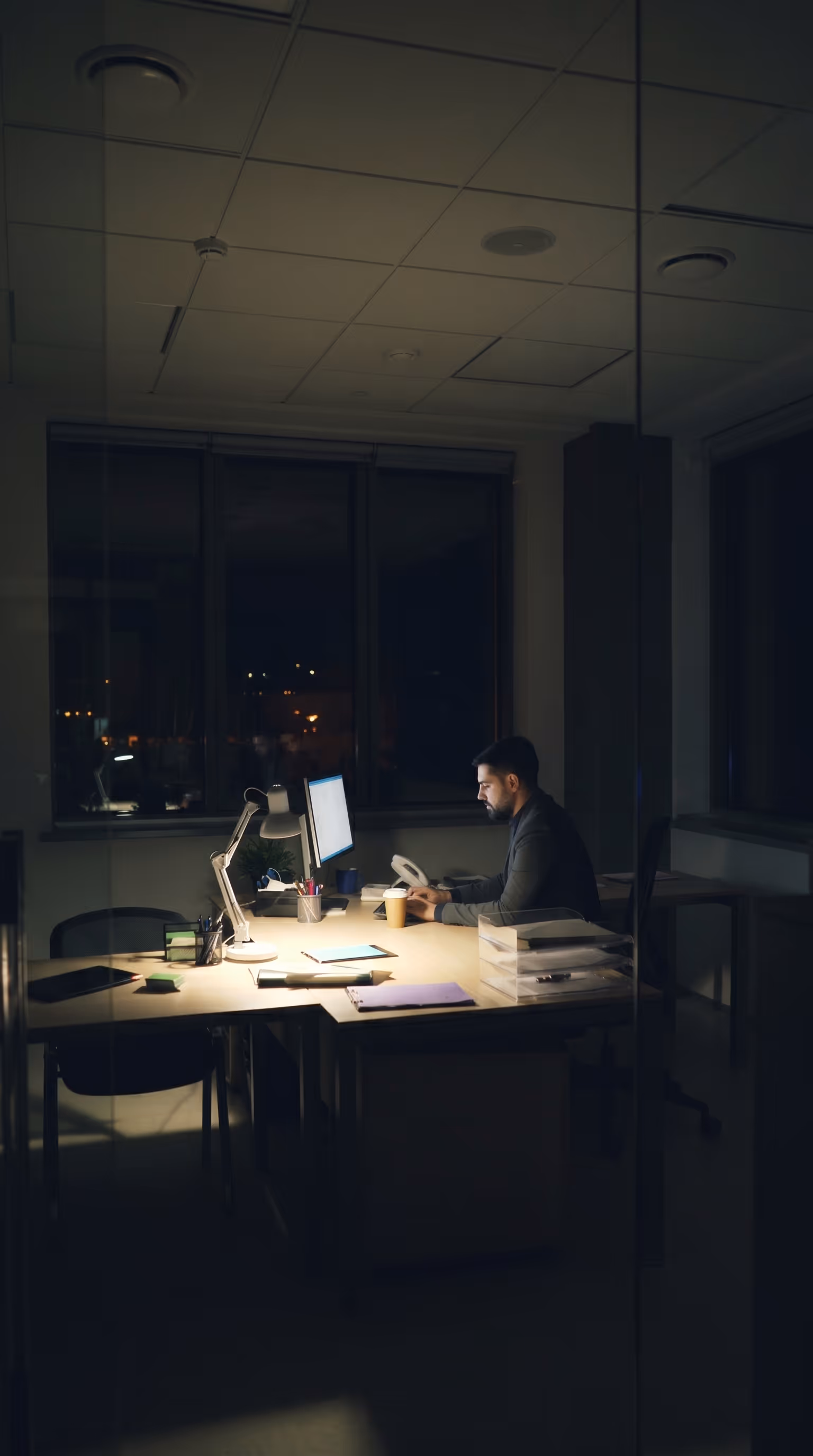 A person working alone at a desk in a dark office late at night, surrounded by documents and lit only by a desk lamp and computer screen, representing the challenge West Country Energy Services faced before Matidor when managers and foremen were reconciling field documentation past 2 a.m. due to aging GIS software and manual data processes.