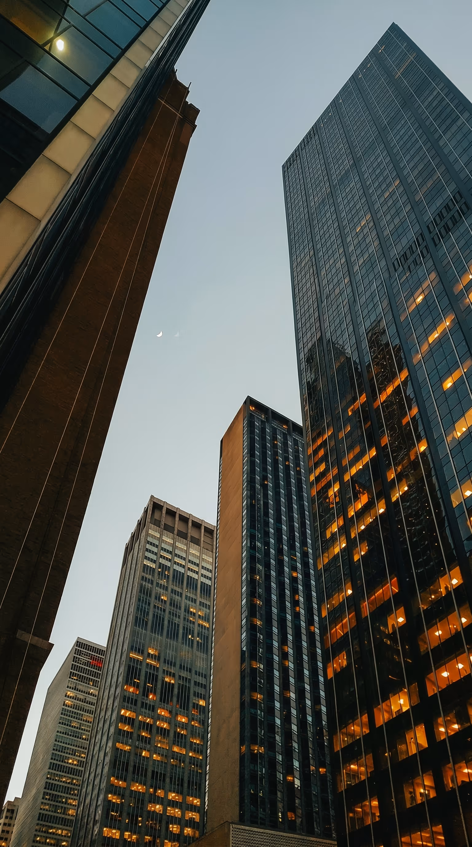 Low-angle view looking up between illuminated glass office towers at dusk with a crescent moon in the sky, representing NorthWind Land Resources Inc., an environmental consulting firm headquartered in Edmonton, Alberta with offices across the province serving oil and gas, land reclamation, and environmental assessment clients.
