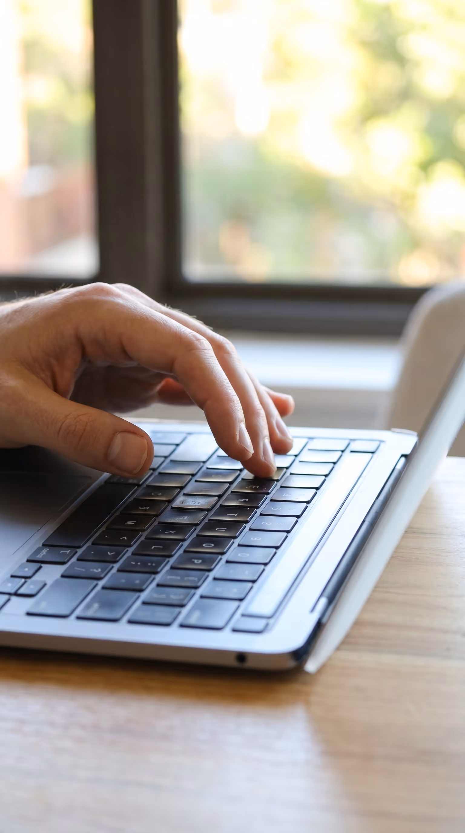 Close up of a hand typing on a laptop keyboard beside a bright window with trees visible outside, representing Tree Time staff updating Matidor field forms like Monitoring Assessments and Inspections directly from site visits to record tree level data and photos for Project Forest.