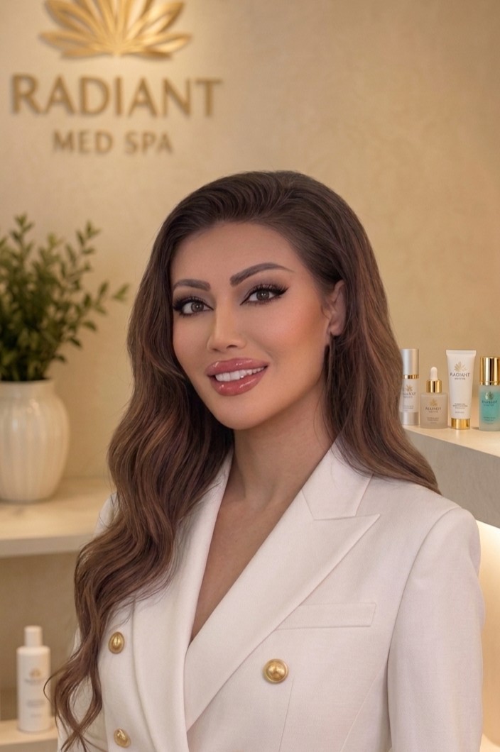 Woman with long wavy brown hair wearing a white blazer with gold buttons smiling in a spa environment with skincare products and a plant in the background.