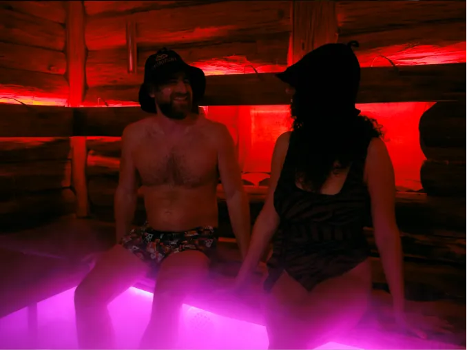 A man and woman sitting happily inside a wooden sauna lit with red and purple lights, both wearing hats and swimwear.