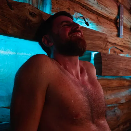 Shirtless man with a beard leaning back against a wooden wall with blue lighting, eyes closed and smiling.