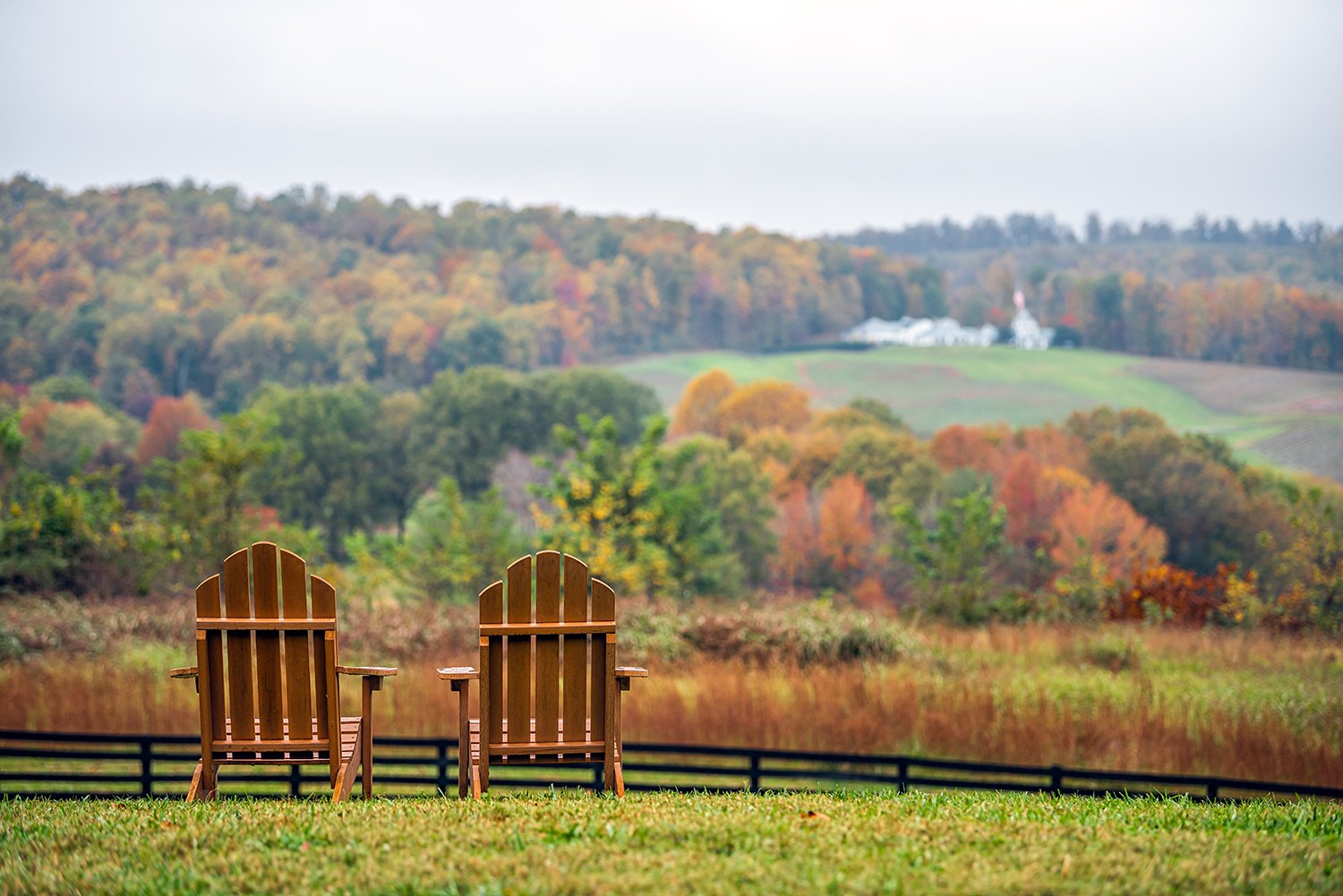 Two wooden Adirondack chairs on green grass facing a scenic autumn landscape with colorful trees and hills.