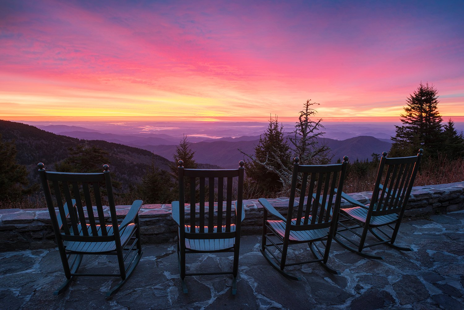 Four empty wooden rocking chairs on stone patio overlooking mountains at colorful sunset.