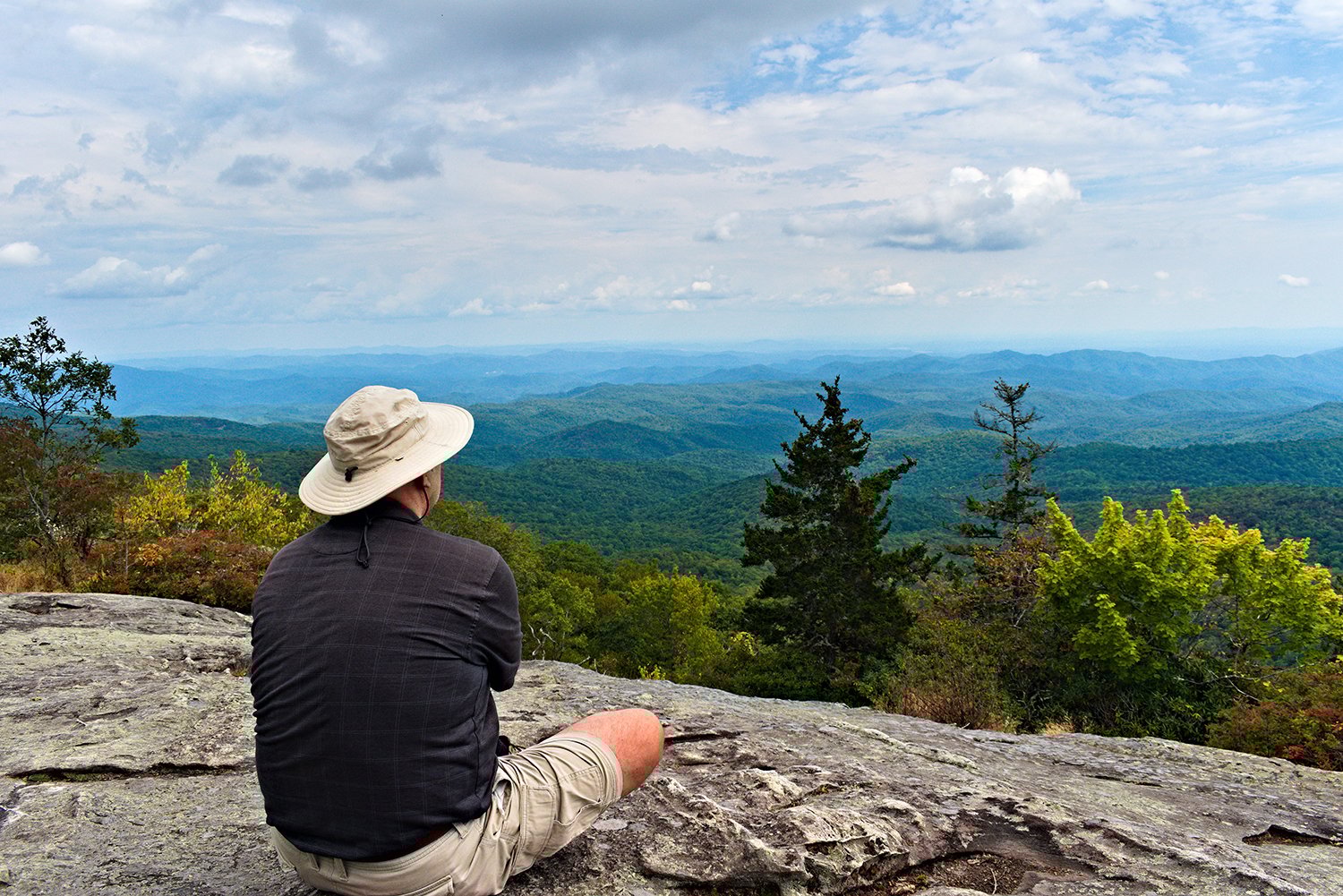Man wearing a wide-brimmed hat and shorts sitting on a rocky cliff overlooking a vast forested mountain range under a partly cloudy sky.